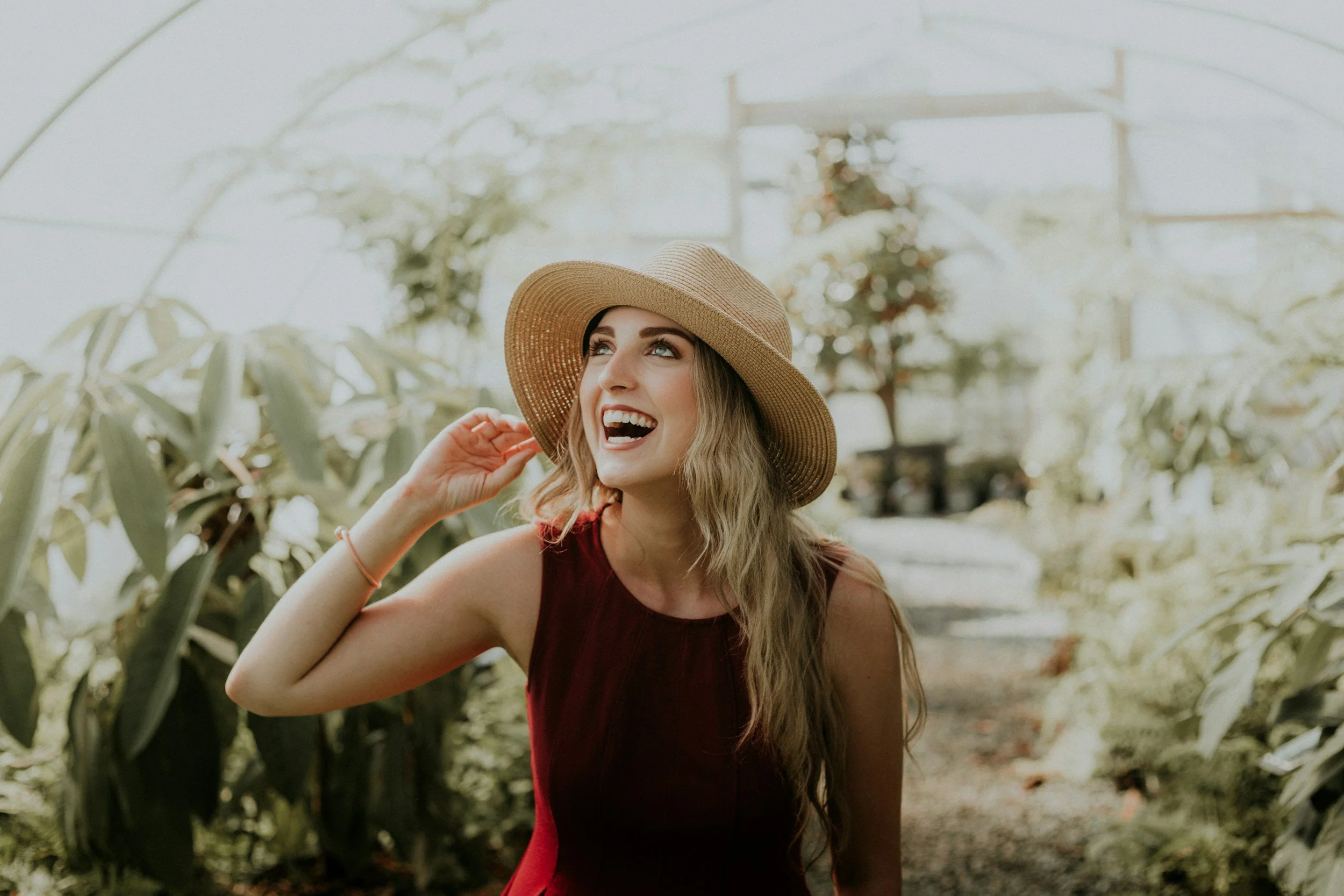 A young woman with long wavy hair wearing a wide-brimmed straw hat and a sleeveless maroon dress, smiling and looking upward inside a greenhouse with lush green plants and trees in the background.