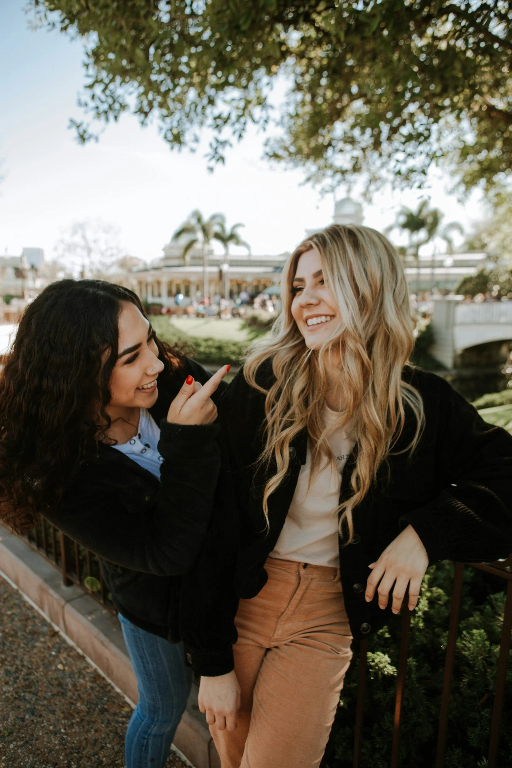 Two women smiling and talking outdoors near a black fence with trees and a building in the background.