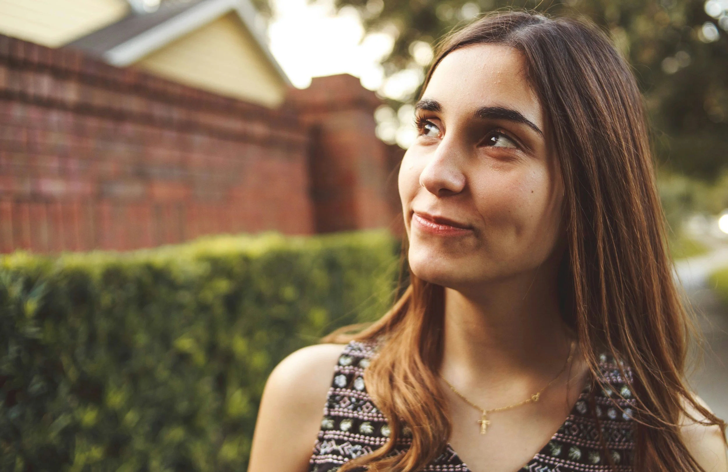 Close-up of a young woman with long brown hair looking to the side outdoors with trees and a brick wall in the background.