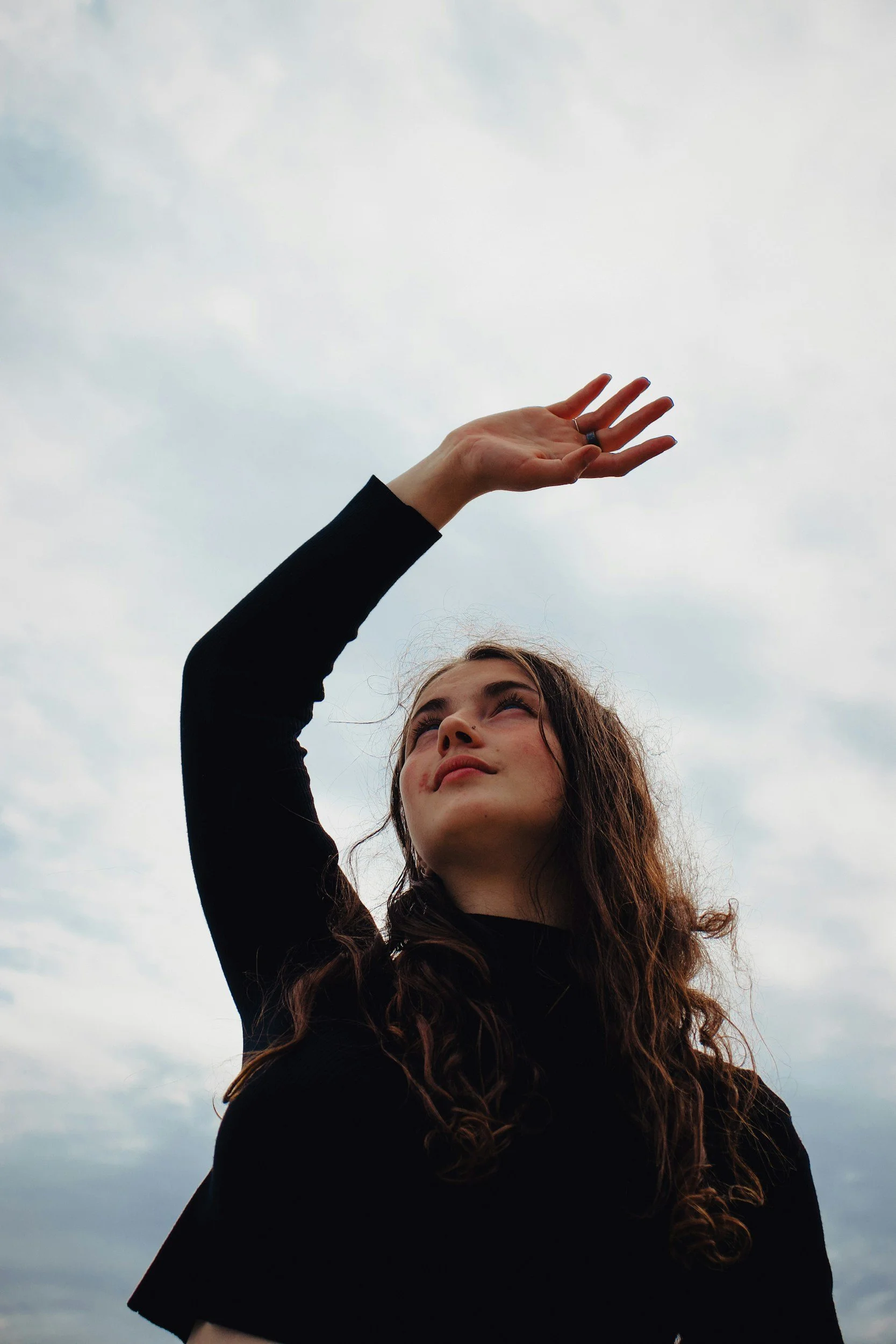 A woman with long brown curly hair looking up and reaching out towards the sky with clouds in the background.