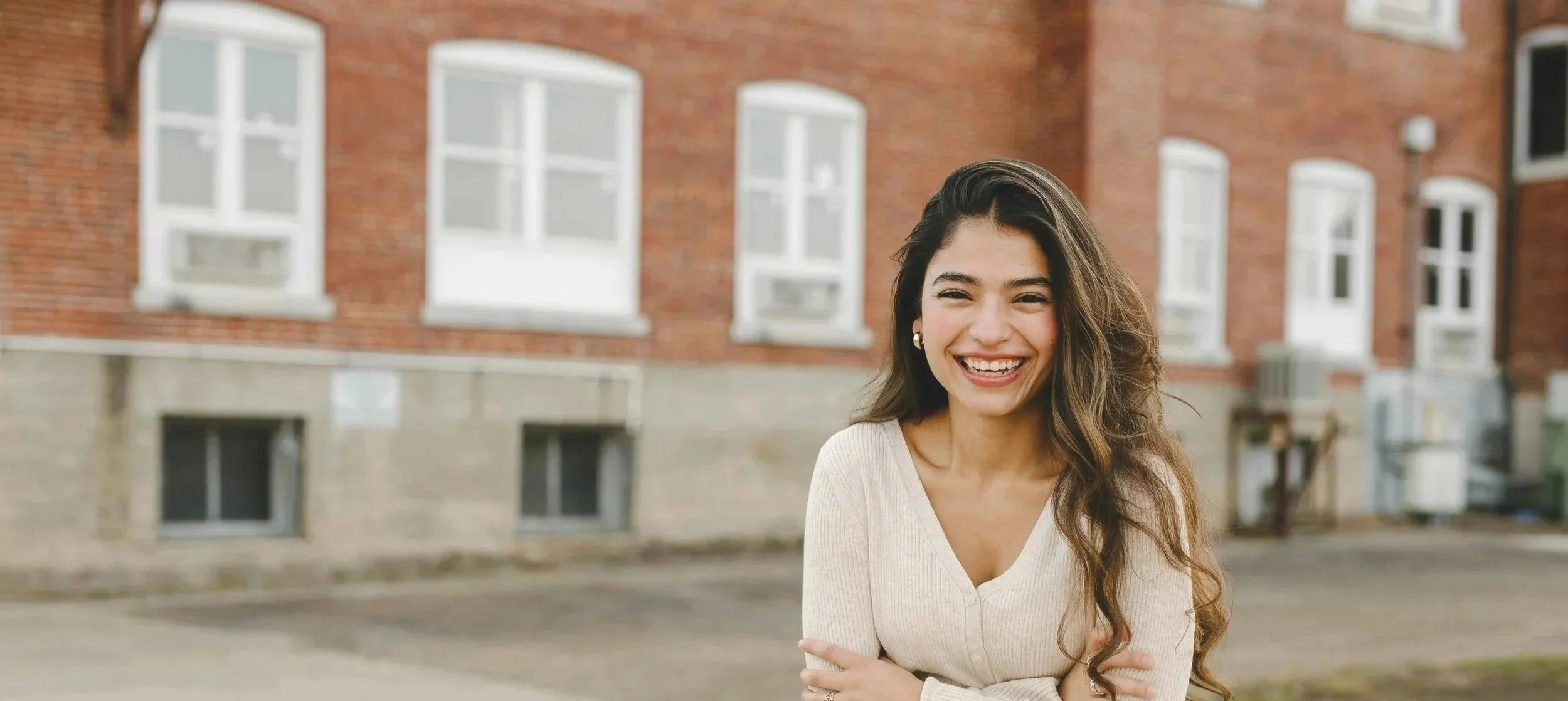 A young woman with long, wavy hair smiling and crossing her arms, standing outdoors in front of a red brick building with multiple windows.