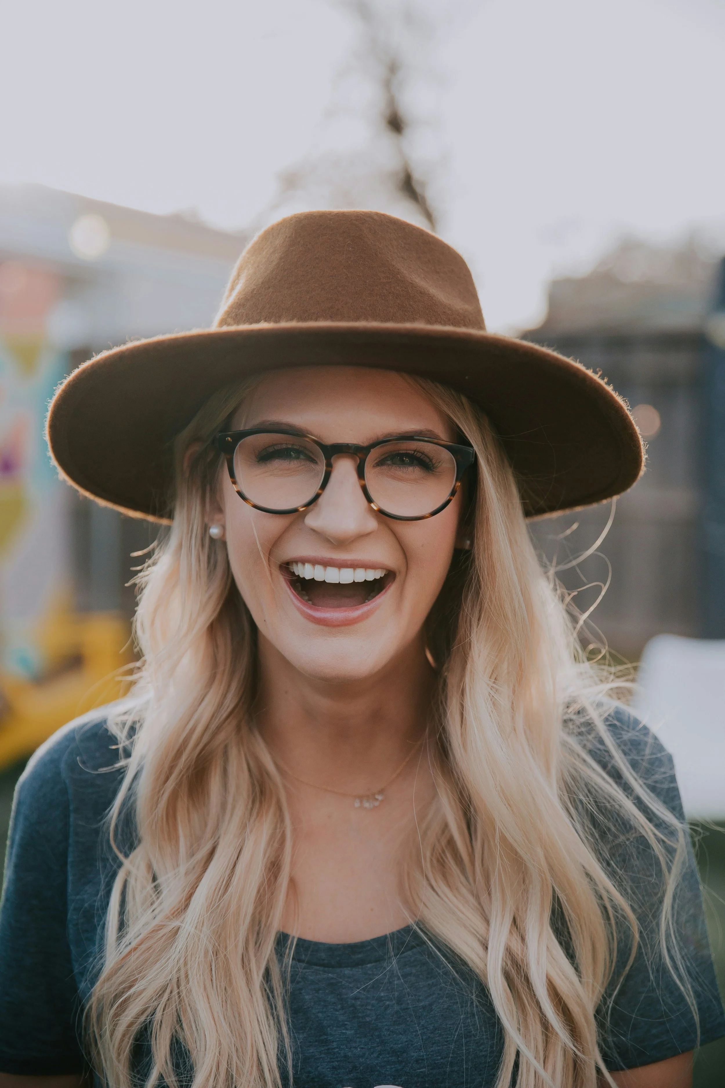 A woman with long blonde hair, wearing glasses, a brown fedora hat, and smiling widely outdoors with buildings and trees in the background.