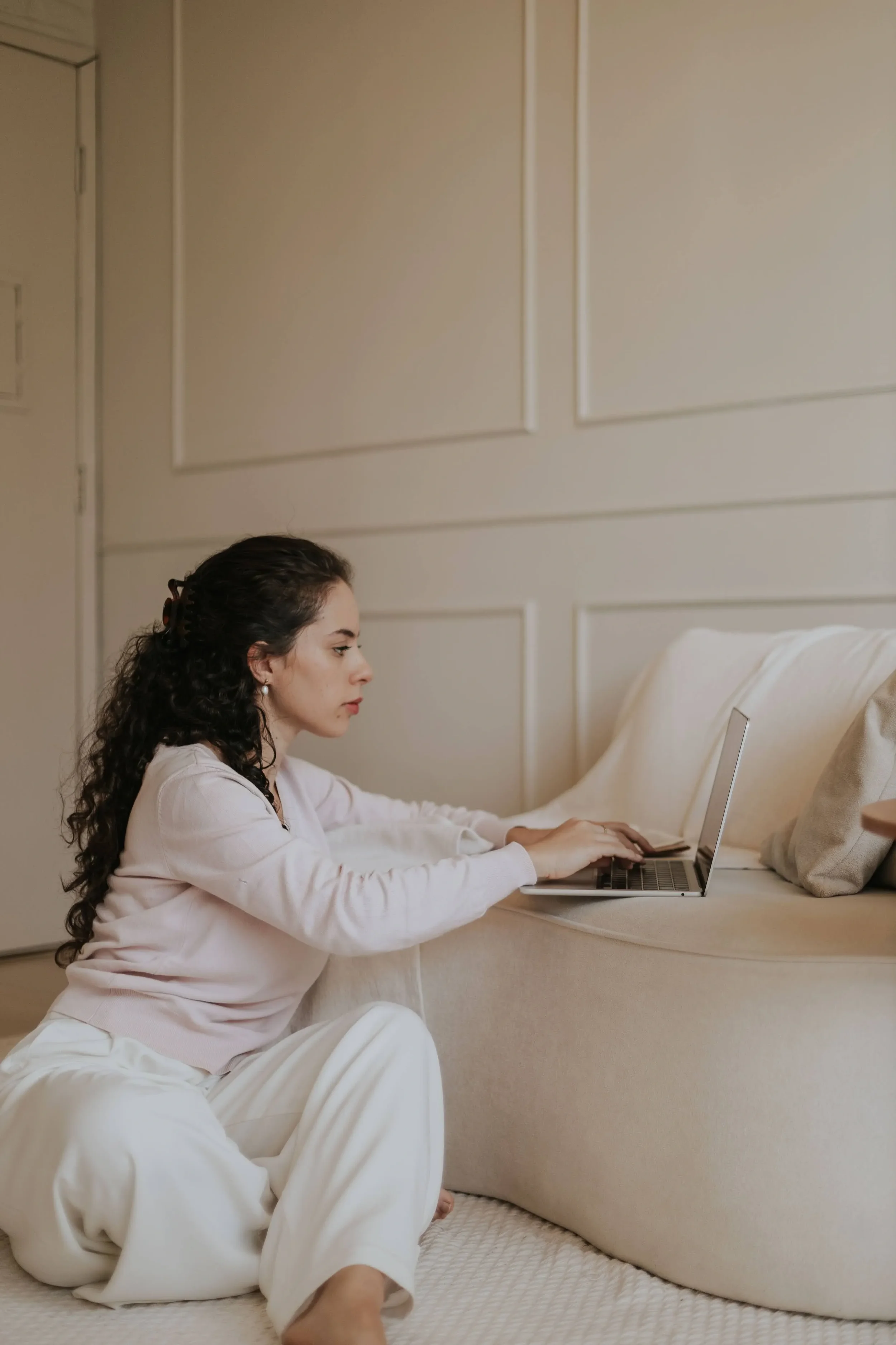 A woman with curly brown hair, dressed in a white robe, is sitting on a bed and working on a laptop. She is in a cozy bedroom with beige walls and a white headboard.