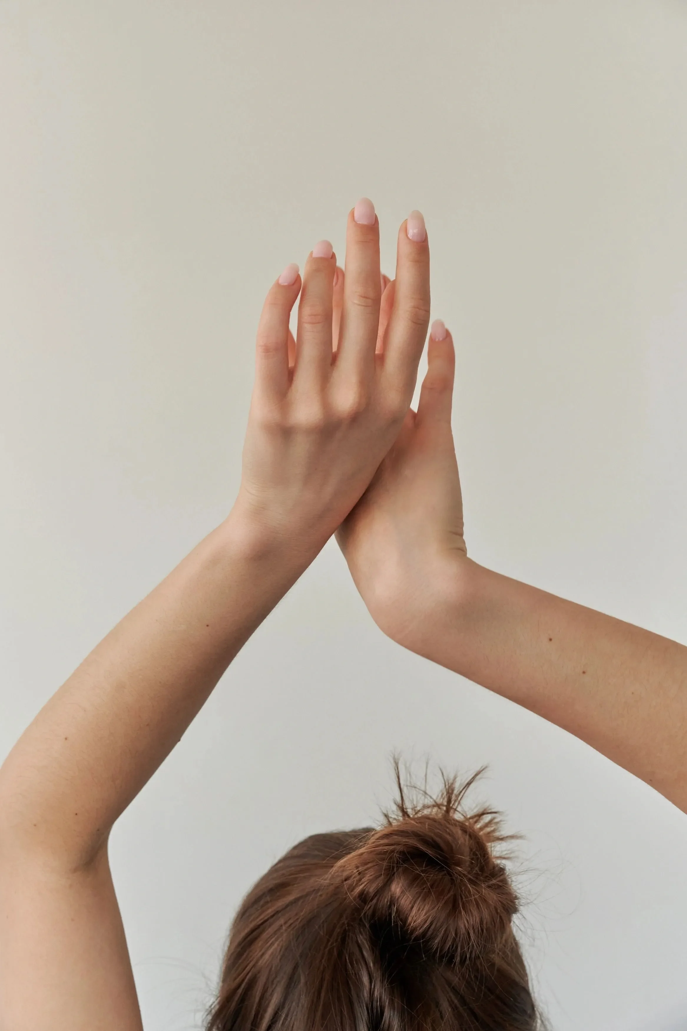 Close-up of a person with reddish-brown hair, head tilted down, and their hands raised above head with one hand overlapping the other, against a light-colored wall background.