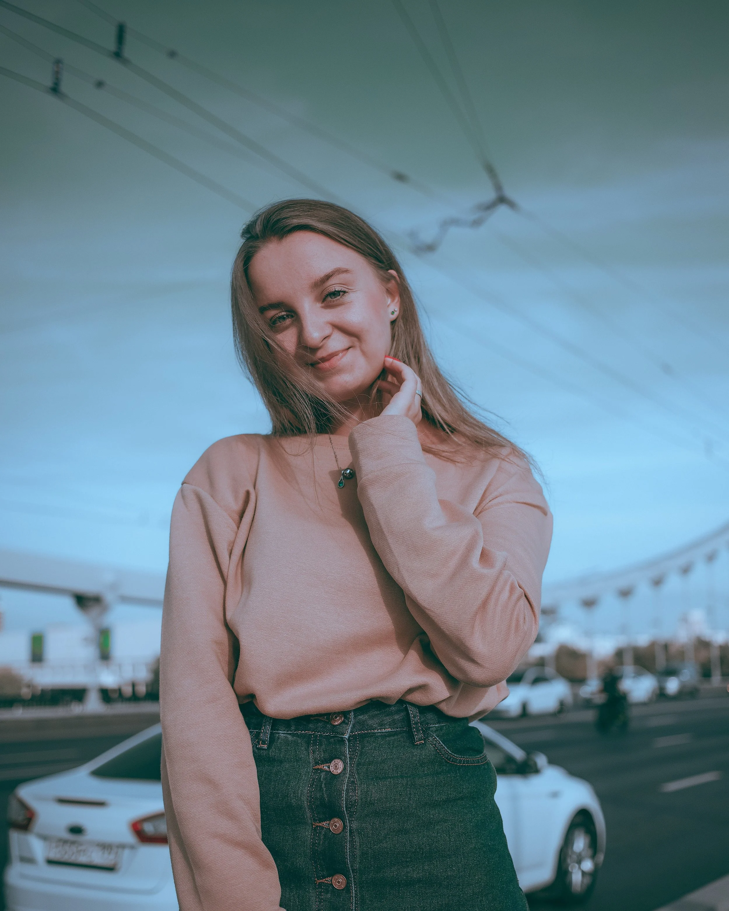 A young woman with long hair, smiling, wearing a beige sweater and dark jeans, standing in front of parked cars in a parking lot, with a structure and sky visible in the background.