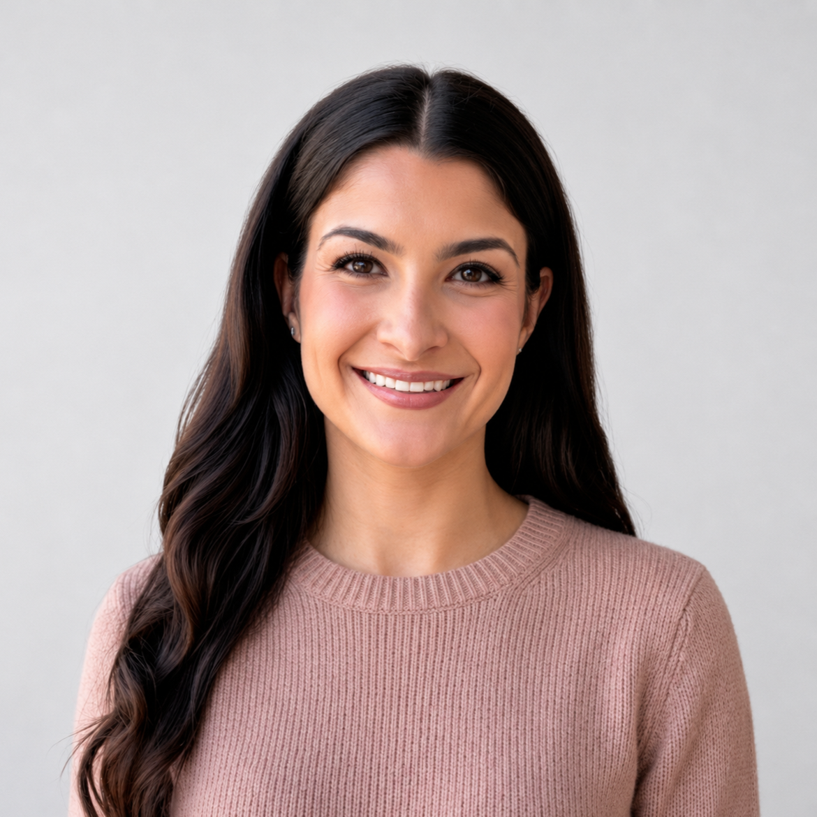 A woman with long dark hair smiling at the camera, wearing a pink sweater, against a plain light gray background.