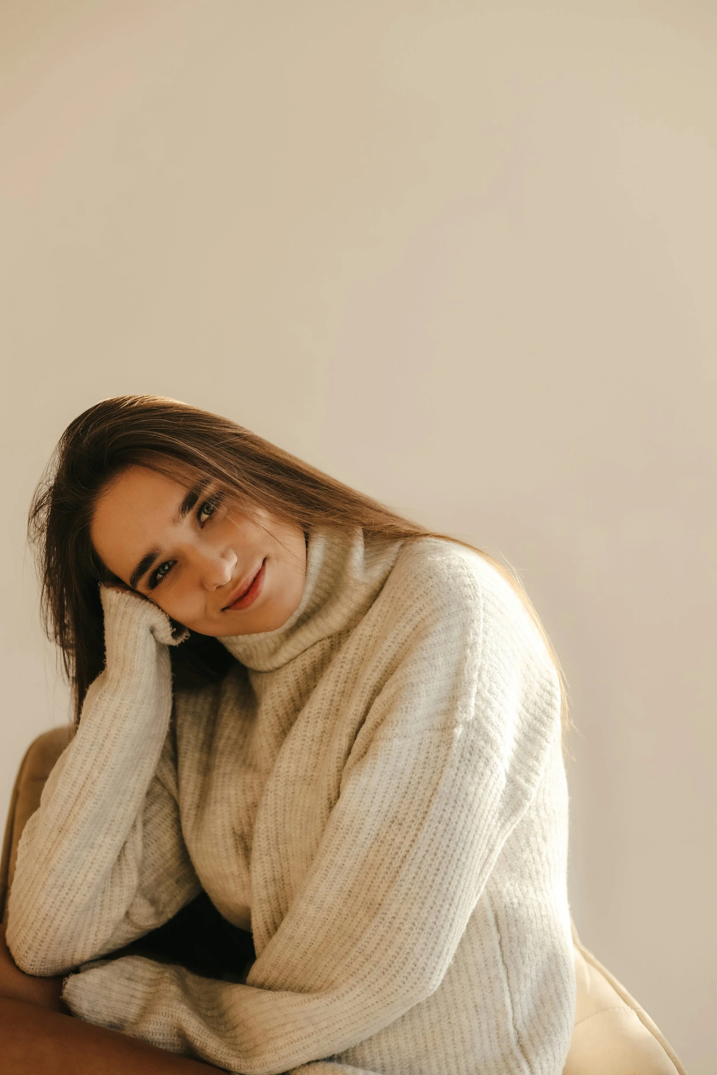 A young woman with long brown hair, wearing a cream-colored knitted sweater, sitting on a chair, smiling gently with her head resting on her hand, against a plain beige wall.