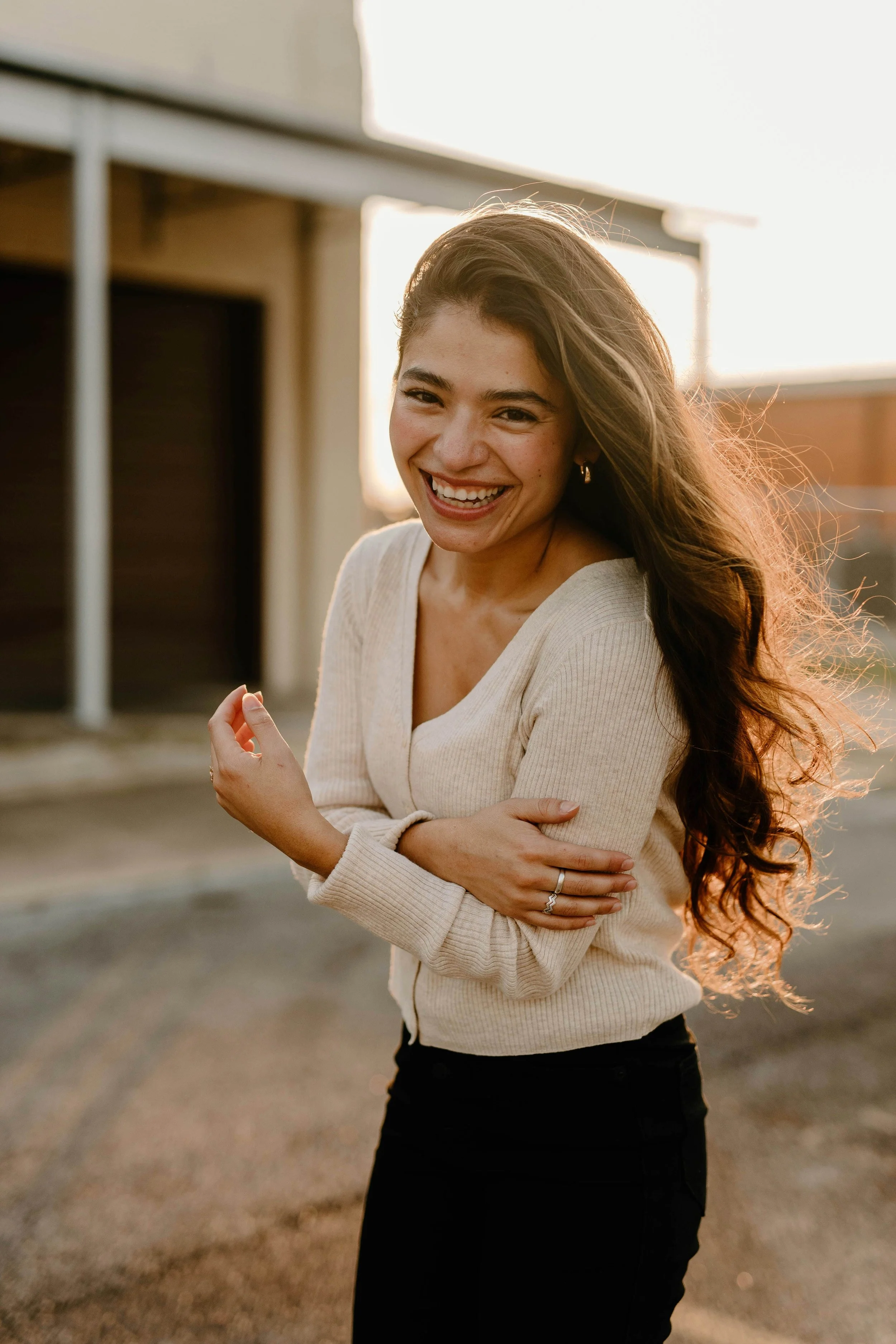 A woman with long, wavy brown hair smiling and looking at the camera, standing outdoors during sunset, wearing a beige sweater and black pants.