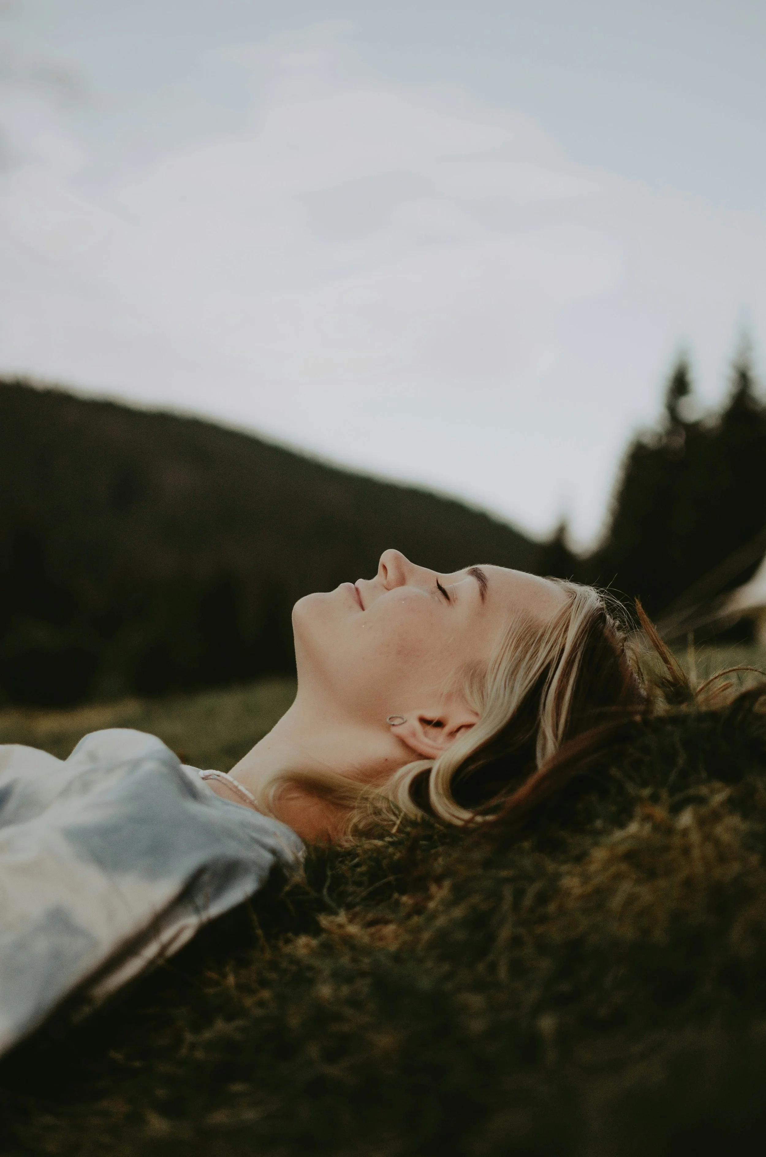 A woman with blonde hair lying on the ground outdoors with her eyes closed, surrounded by green hills and trees under a cloudy sky.
