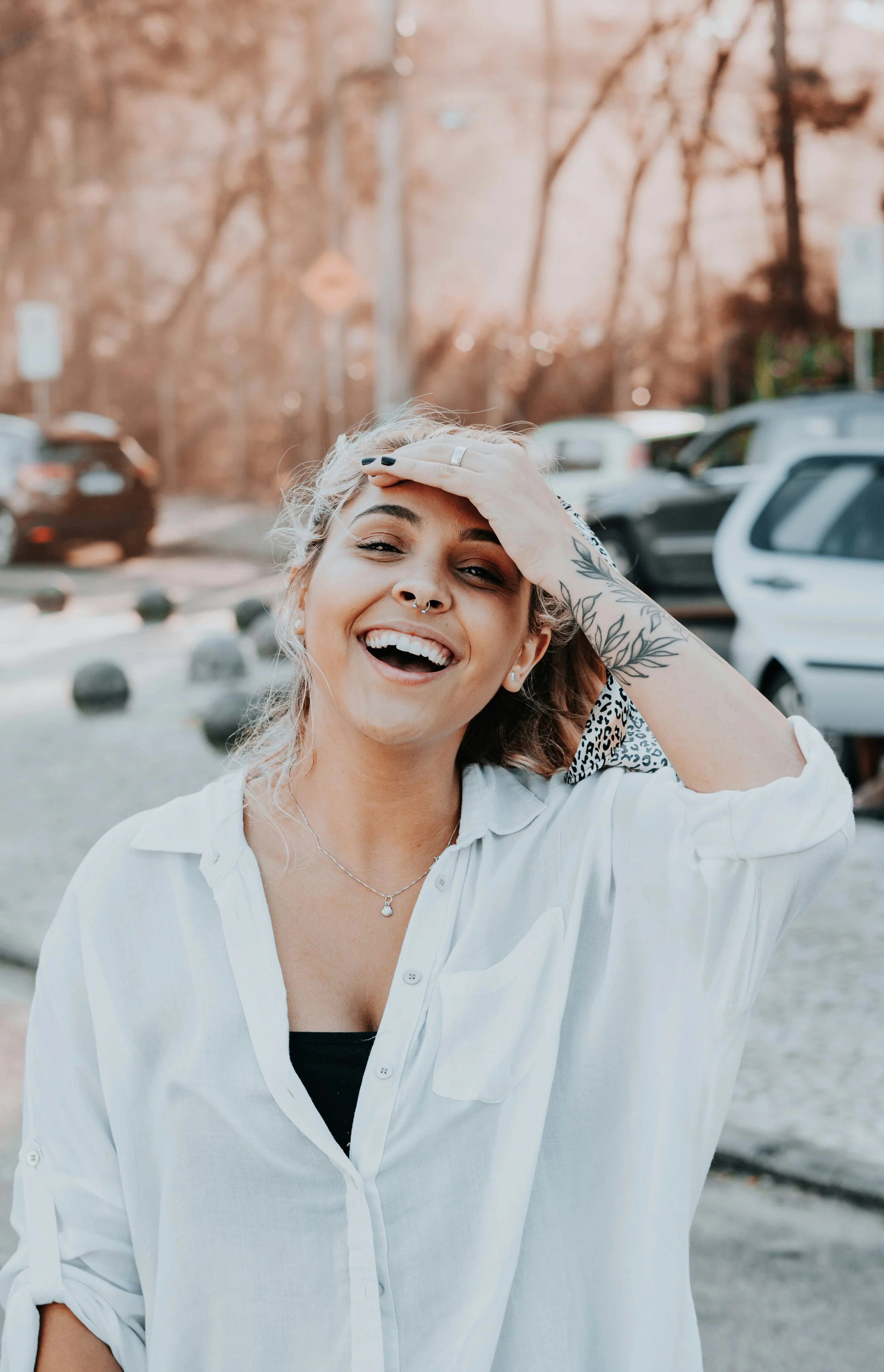 A woman with blonde hair, tattoos on her arm, and a septum piercing smiling outdoors in a parking lot, touching her forehead with her hand.