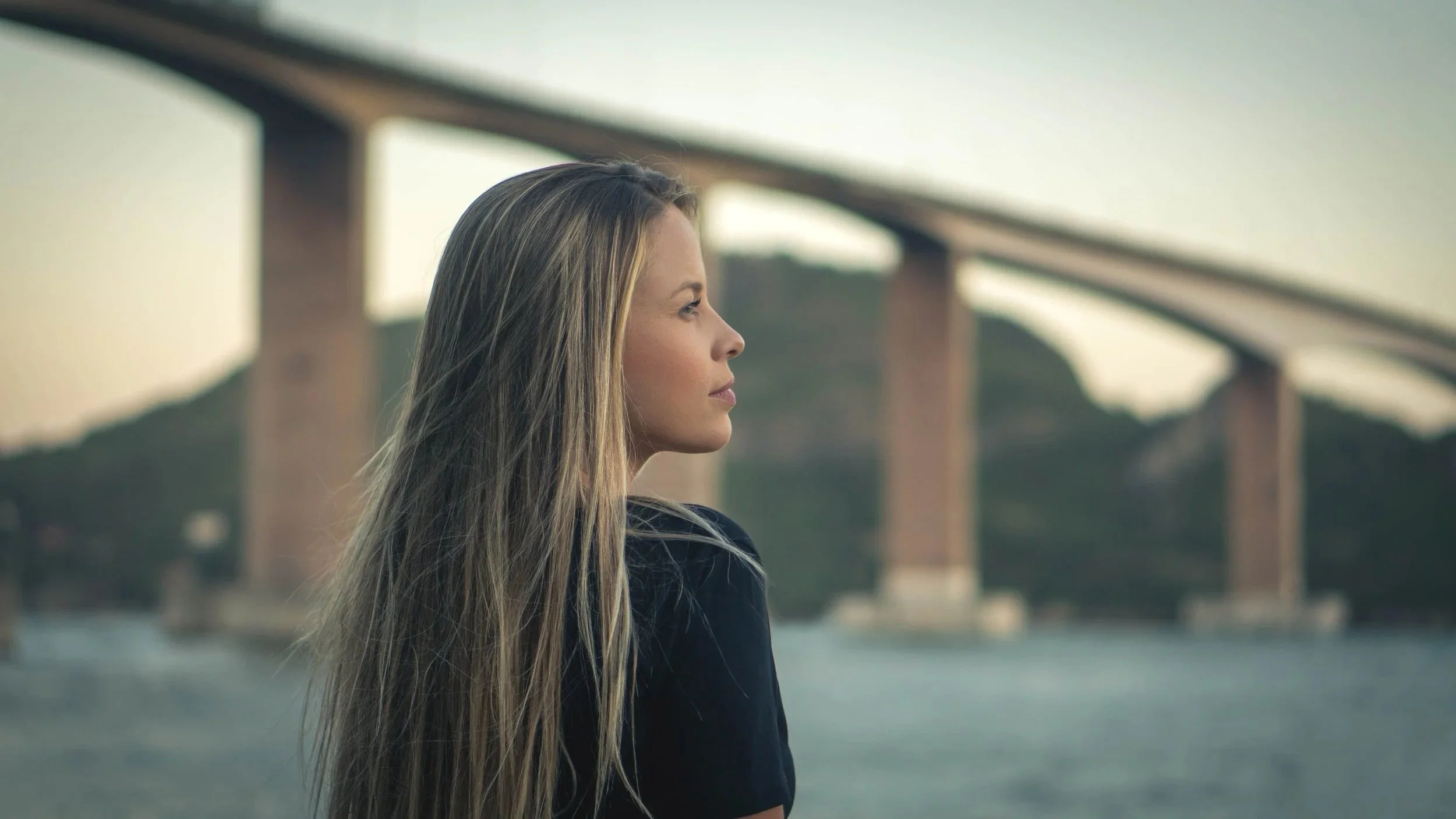Side profile of a woman with long blonde hair standing outdoors near a body of water with a bridge in the background during daytime.