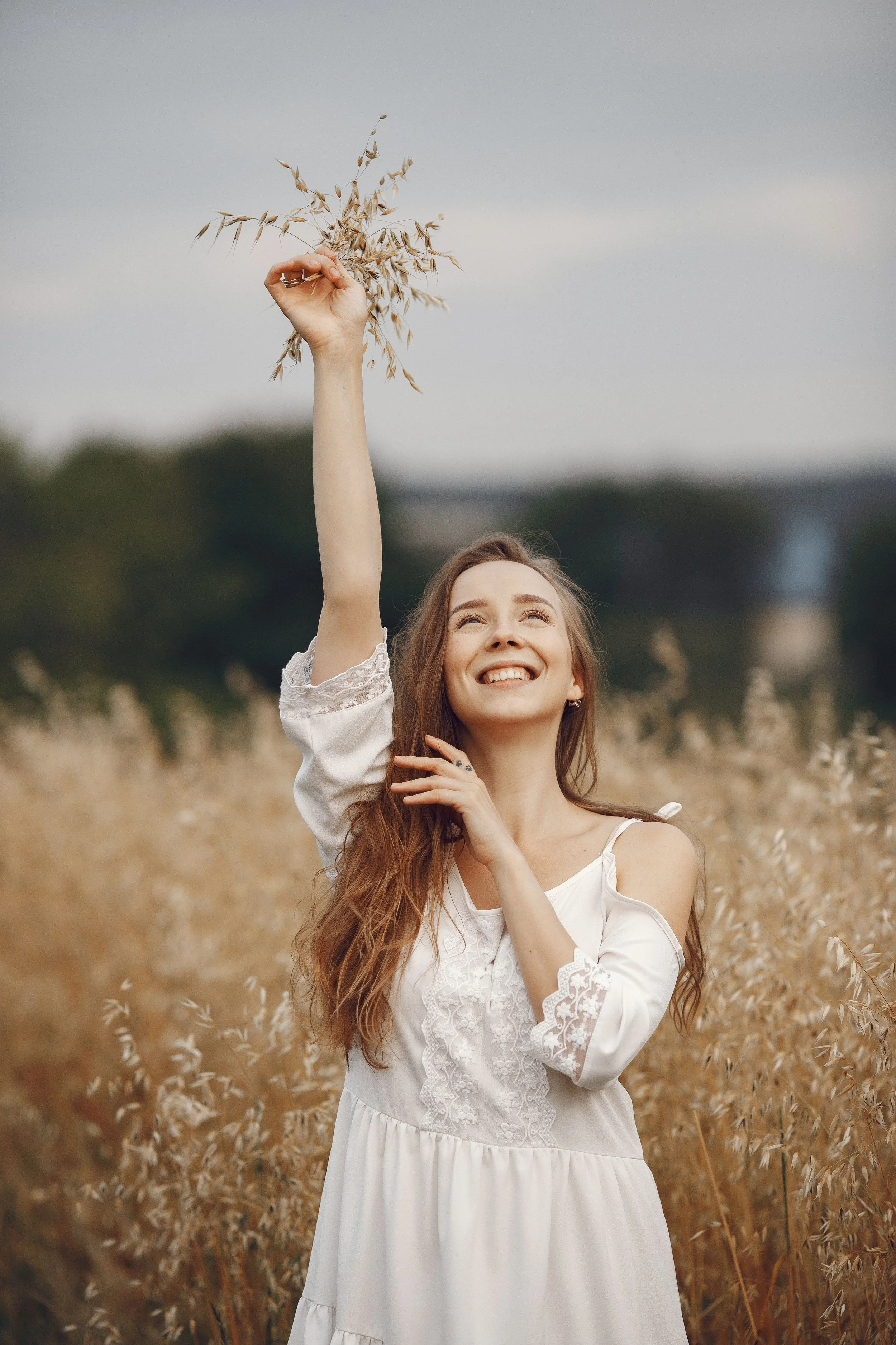 A woman with long red hair in a white dress standing in a golden field of tall grass, smiling and holding a dried plant against a cloudy sky.