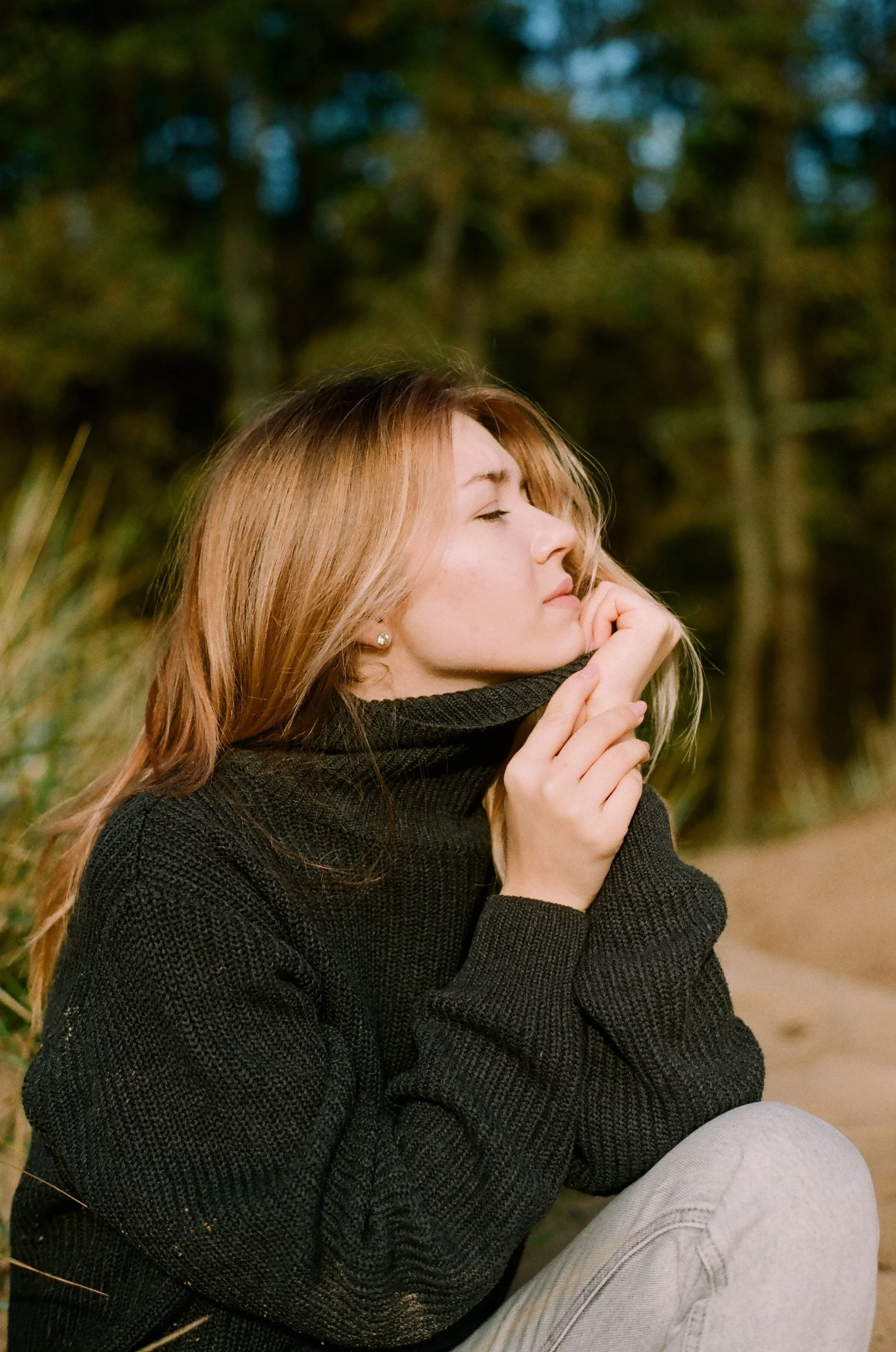 A young woman with red hair, wearing a black sweater, sitting outdoors with her eyes closed and hands clasped near her face, surrounded by greenery.