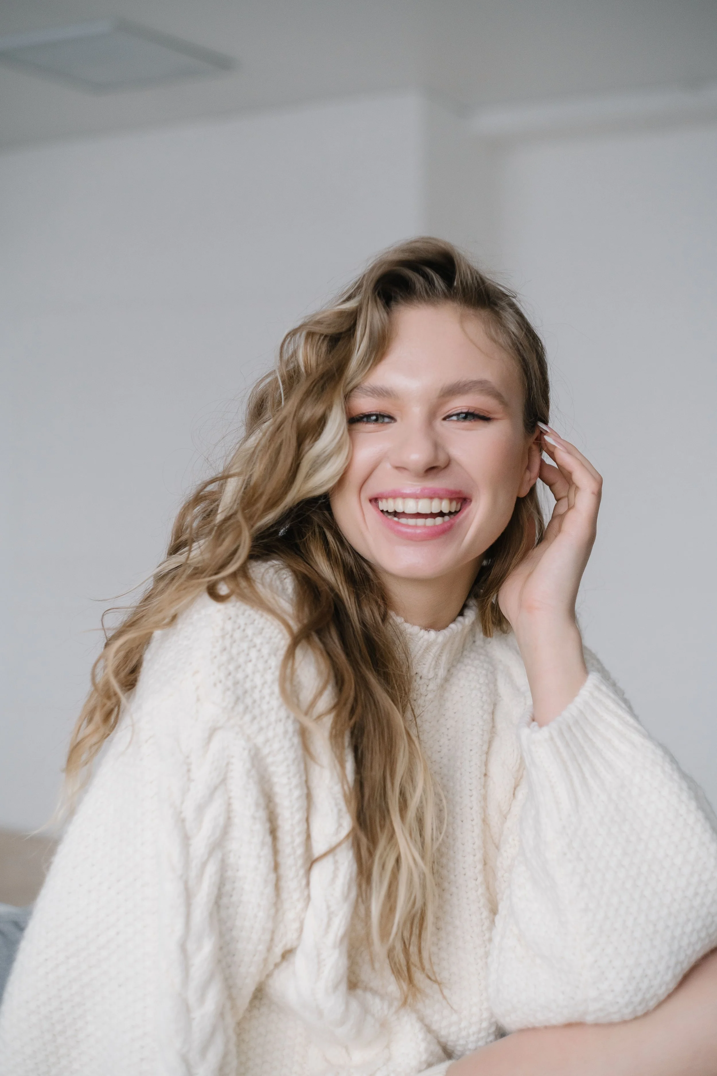 A young woman with long curly blonde hair smiling and looking at the camera, wearing a white knit sweater and sitting in a room with a plain white wall.