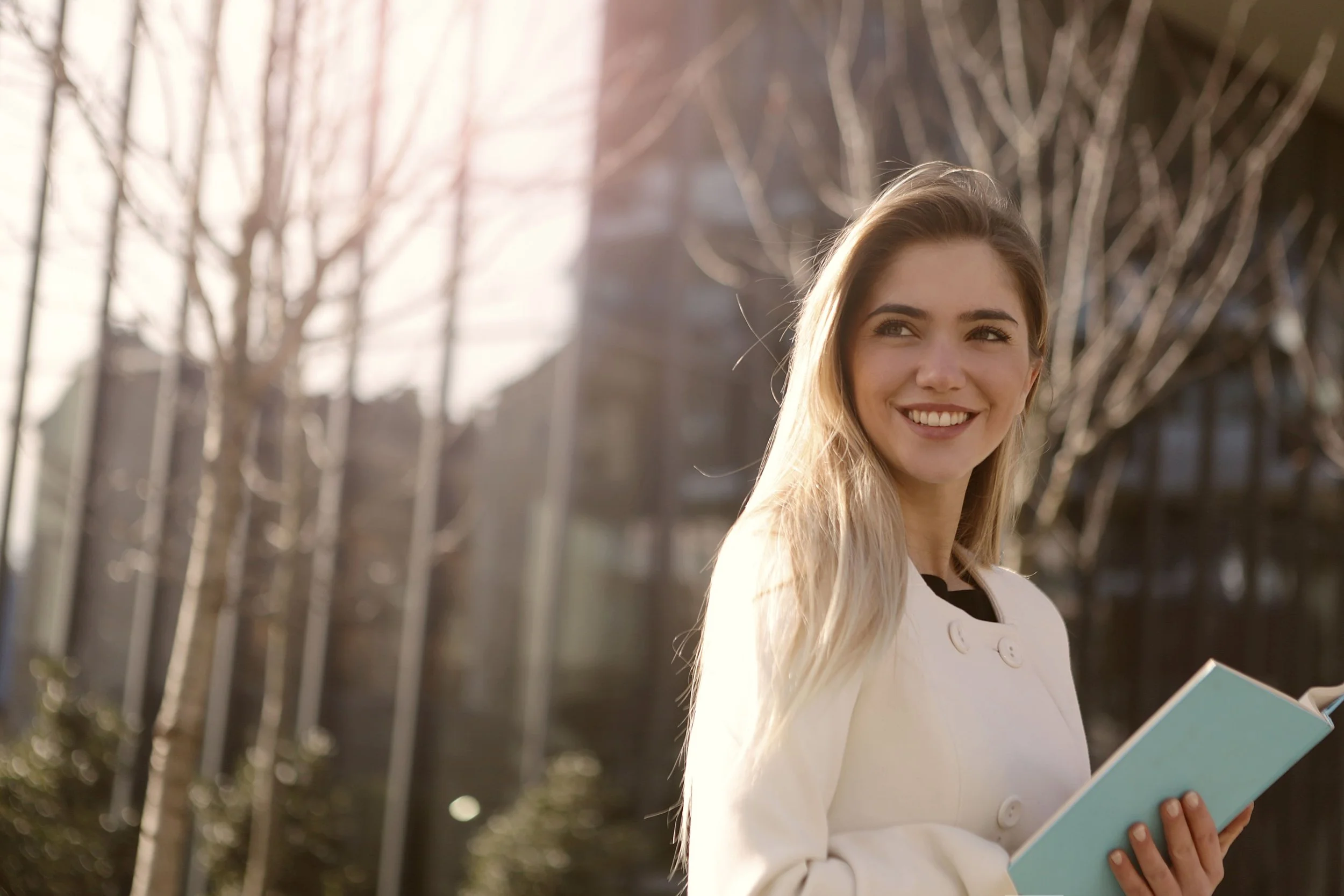 A young woman with long blonde hair smiling outdoors, holding a light blue book.