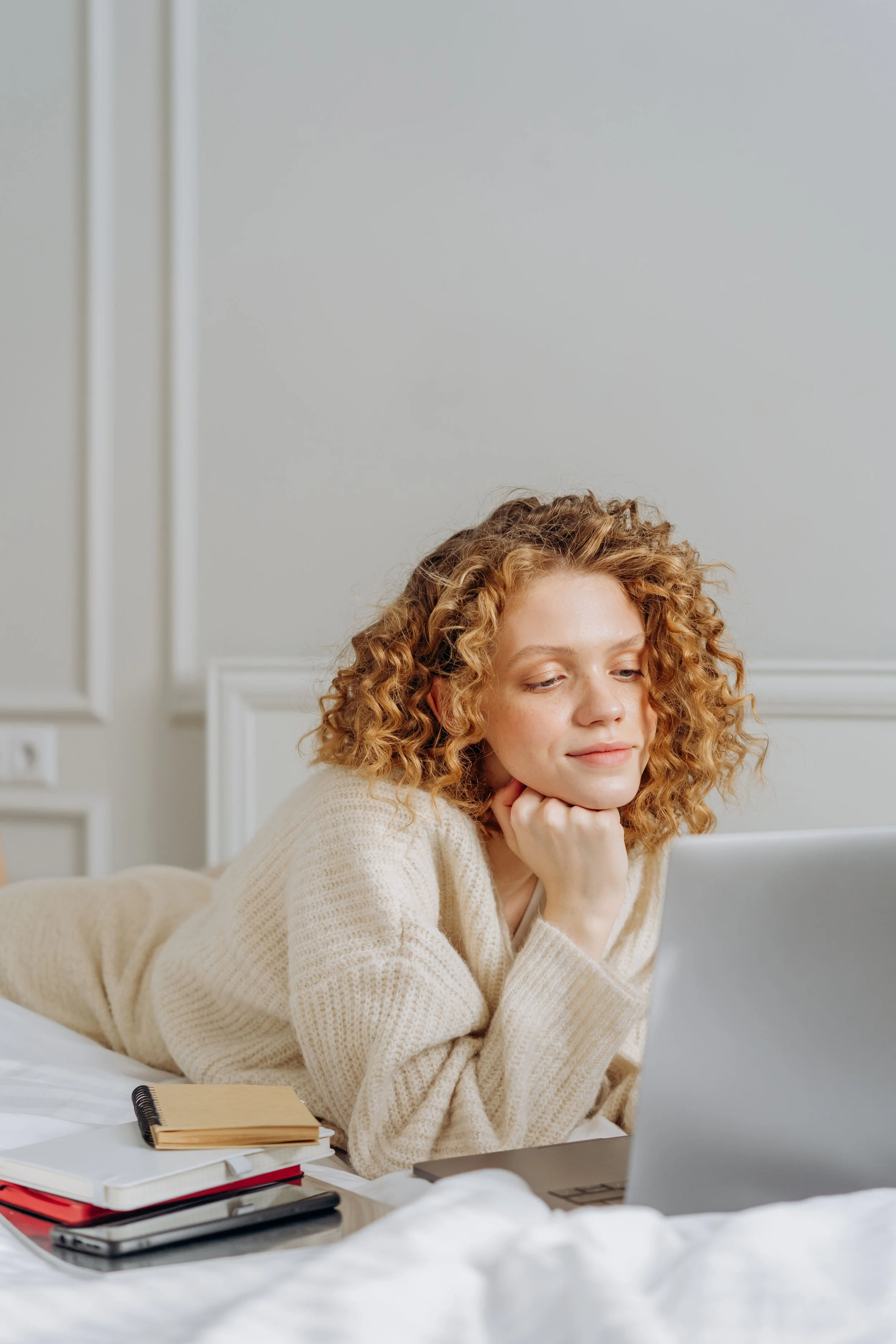 Woman with curly red hair lying on her stomach on a bed, looking at a laptop, with notebooks and phone on the bed, in a room with white walls.