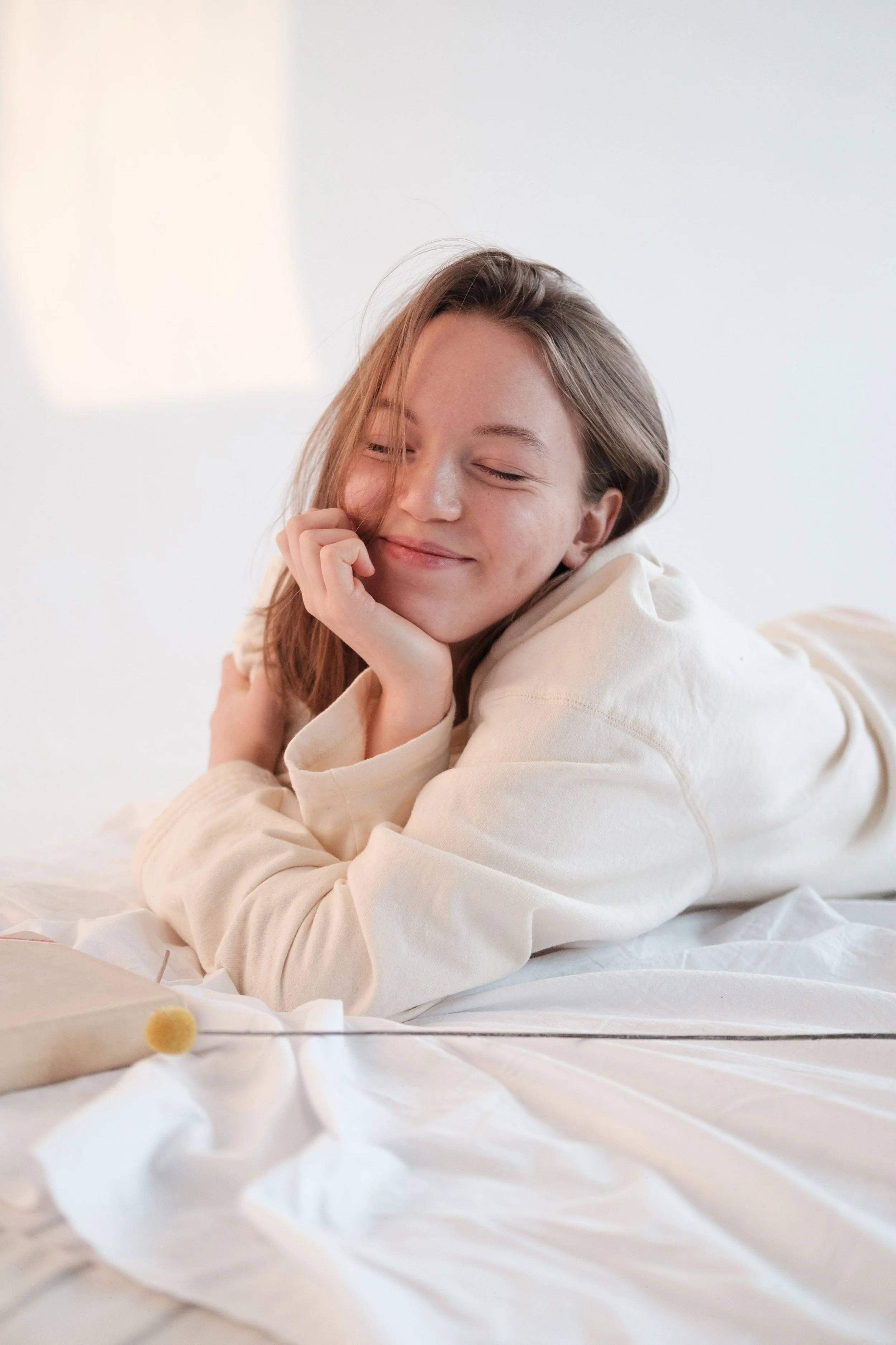 A young woman with long brown hair, eyes closed and a content smile, resting her face on her hand on a bed with white sheets, in a bright room with soft natural lighting.