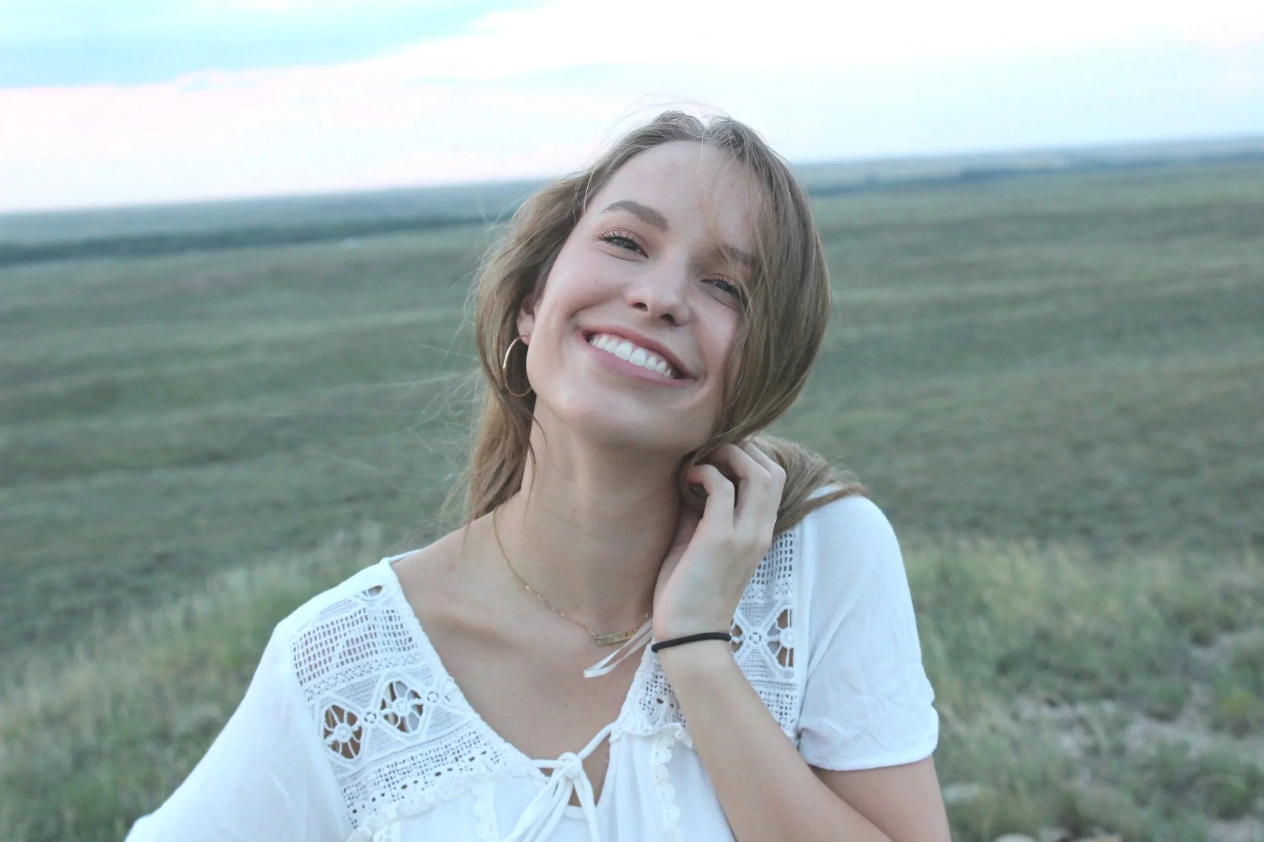 A young woman with light brown hair smiling outdoors in a green field, wearing a white lace top and hoop earrings.