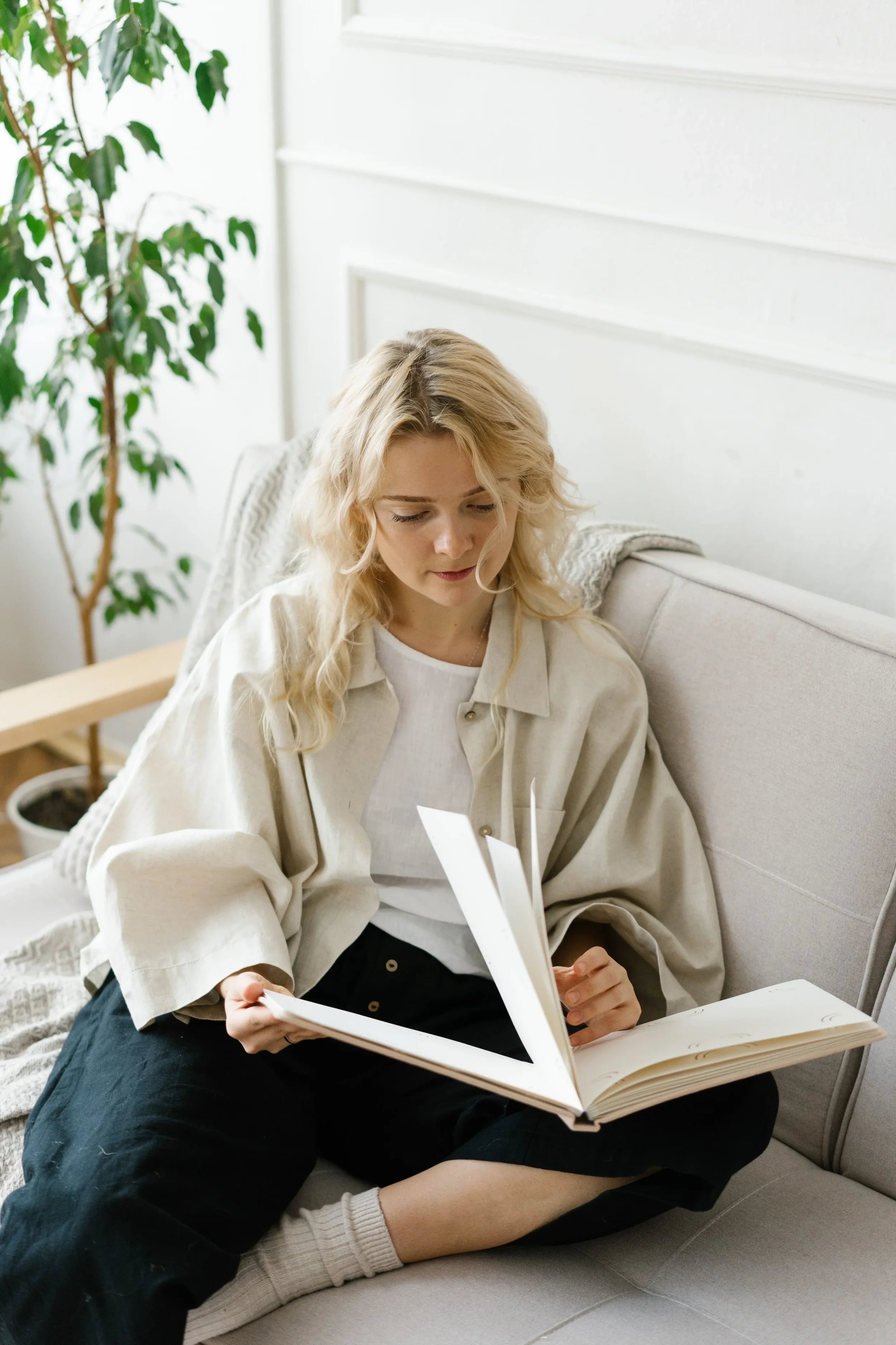 A woman with blond, curly hair sitting on a gray sofa, wearing a cream-colored jacket and black pants, reading a photo album. She is also wearing beige socks and gray shorts. A potted plant with green leaves is in the background.