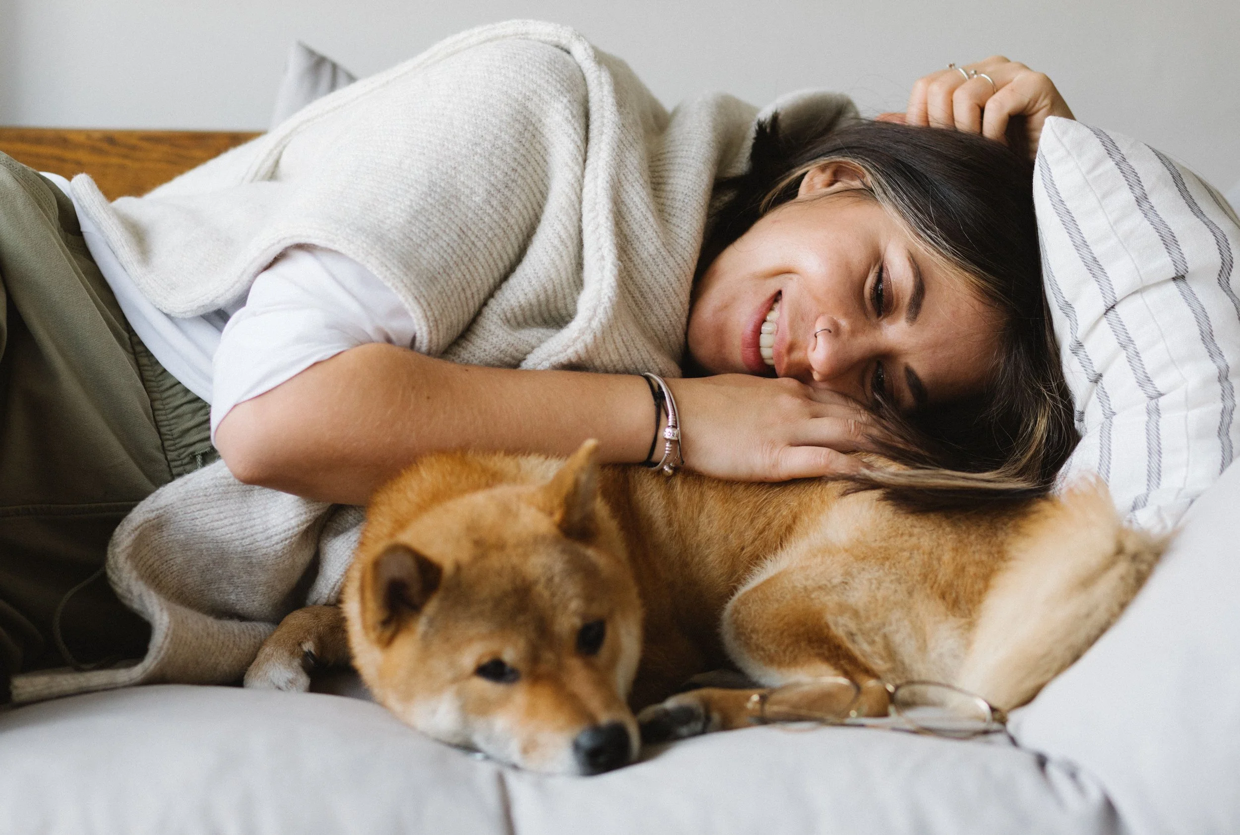 A woman lying on her side on a bed, smiling, with a Shiba Inu dog resting near her, all indoors.