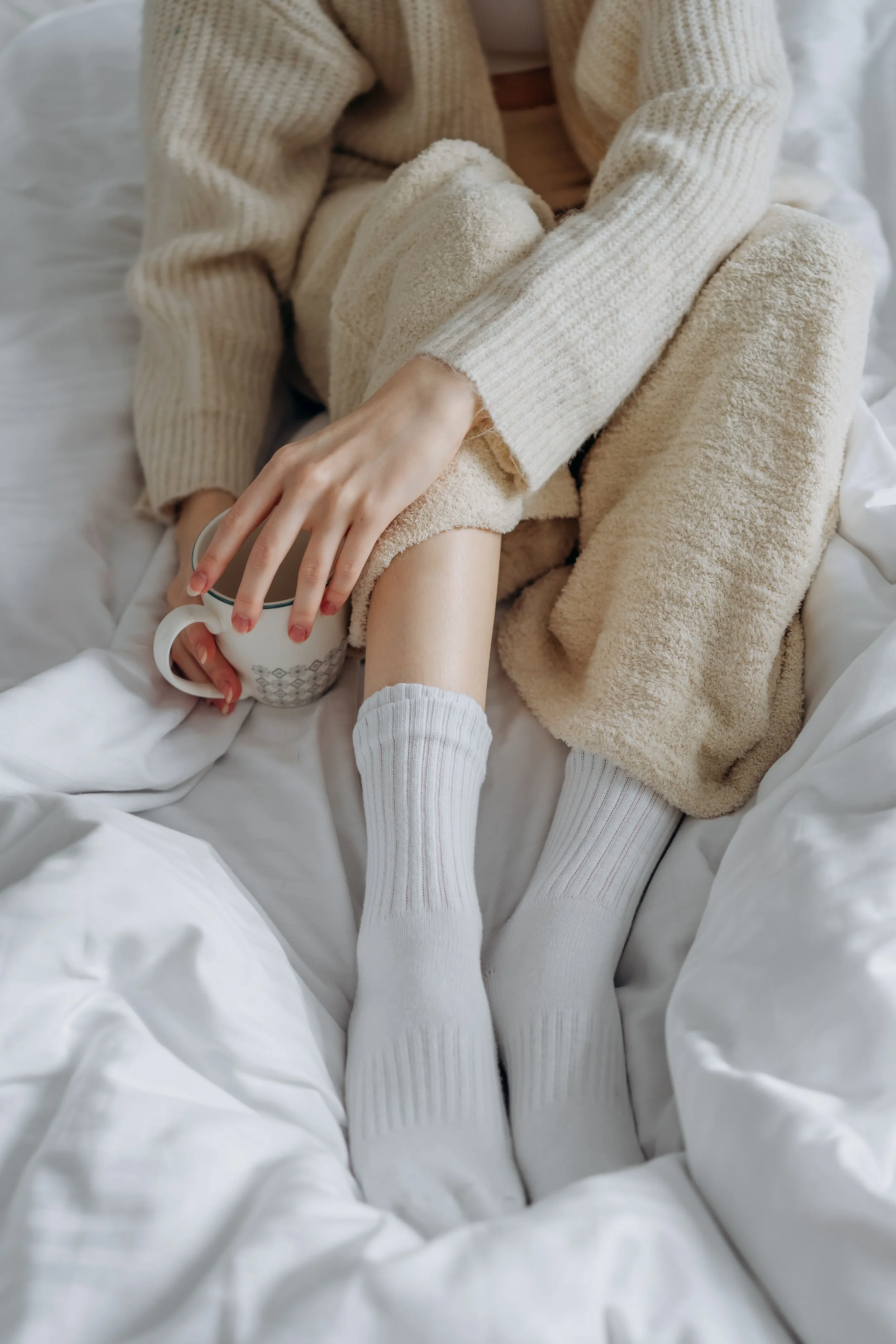 Person wearing knitted beige and white socks, holding a white mug with a pattern, lying on a bed with white sheets, wearing cozy beige and white layered clothing.