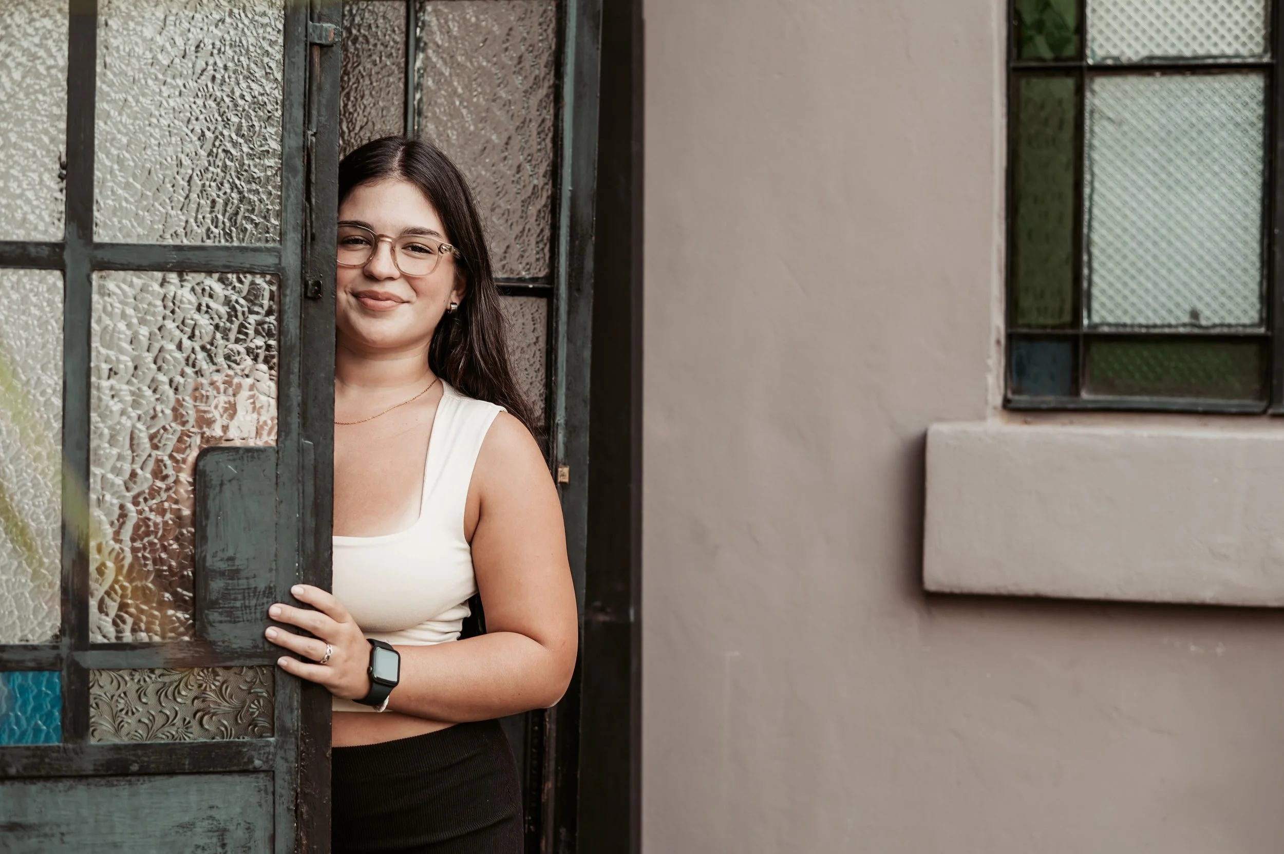 A woman with long dark hair, wearing glasses, a white sleeveless top, and a smartwatch, peeking out from behind a textured glass and metal door, standing next to a window with textured glass on a beige wall.