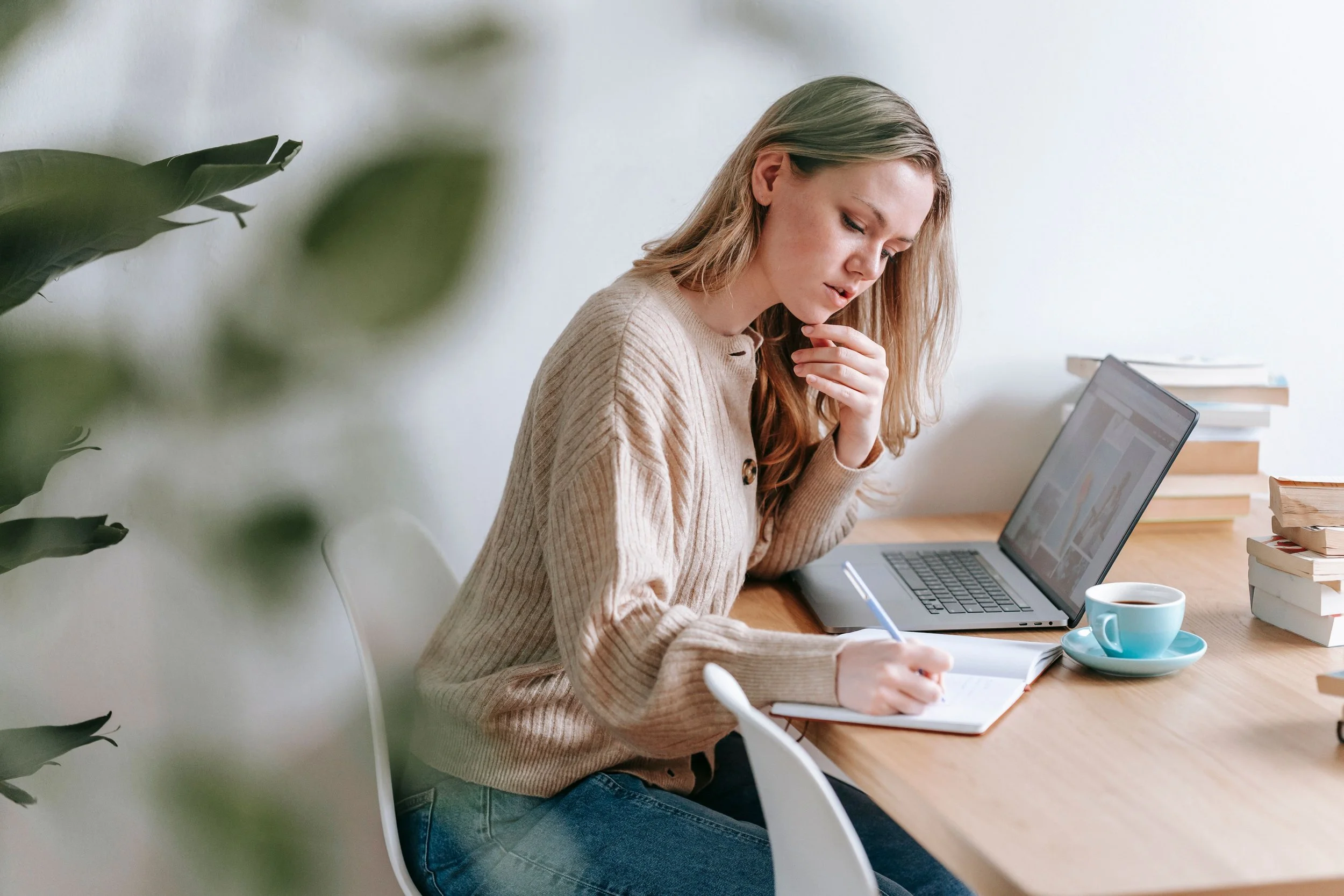 A young woman with long reddish hair is seated at a wooden desk, focused on writing in a notebook. She is wearing a beige sweater and blue jeans. On the desk, there is an open laptop, a blue coffee cup on a saucer, and a stack of books. A blurred leafy plant partially obscures the left side of the image.