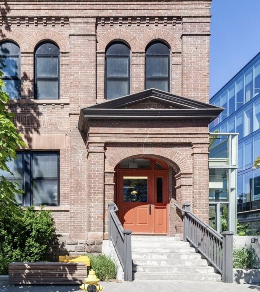 Red brick building with arched windows, and a wood and glass front door accessed by a short flight of stairs with a gray railing. Modern glass building on the right, greenery on the left, and a fire hydrant in front.