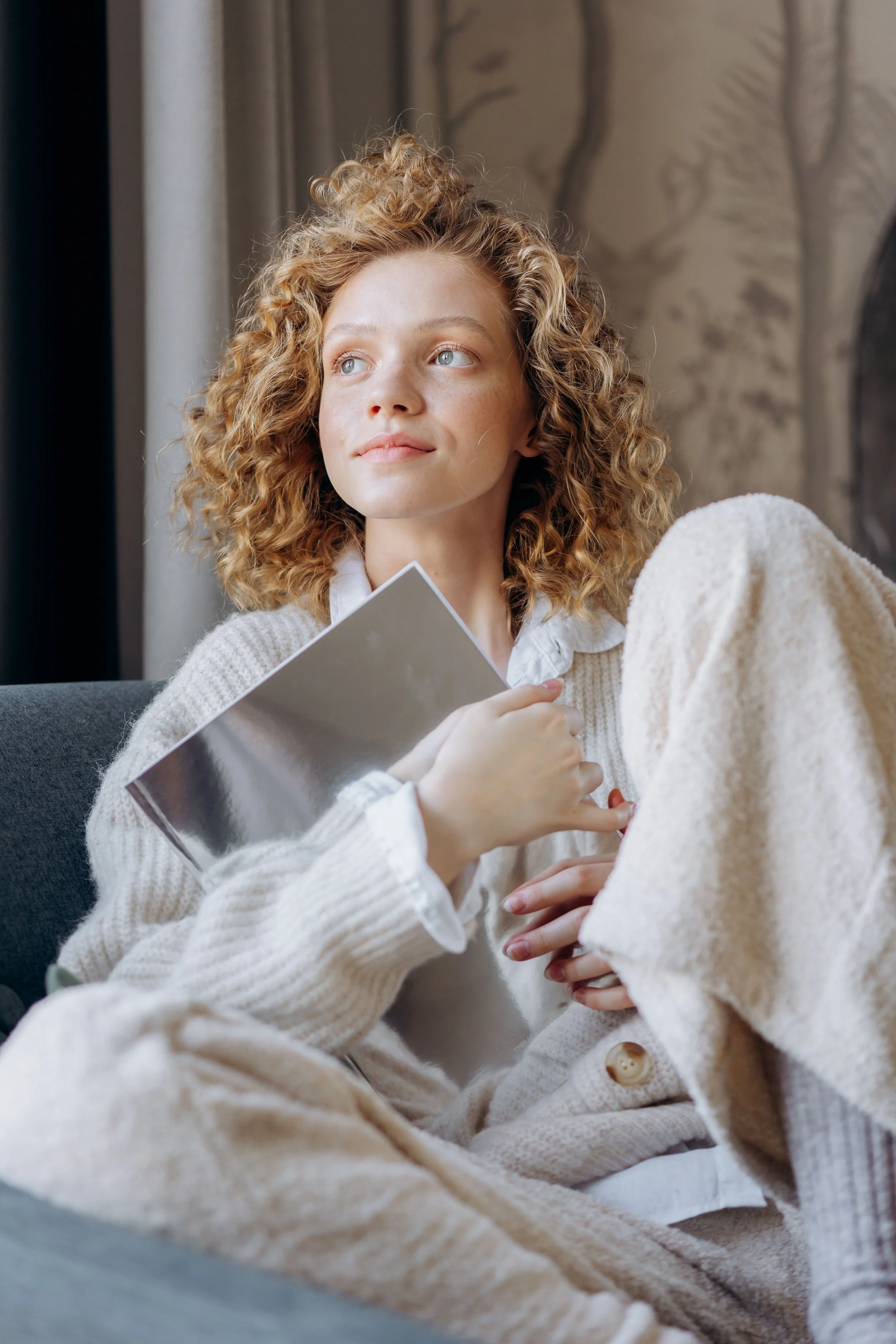 A young woman with curly blonde hair sitting on a couch near a window, holding a book, gazing thoughtfully outside.