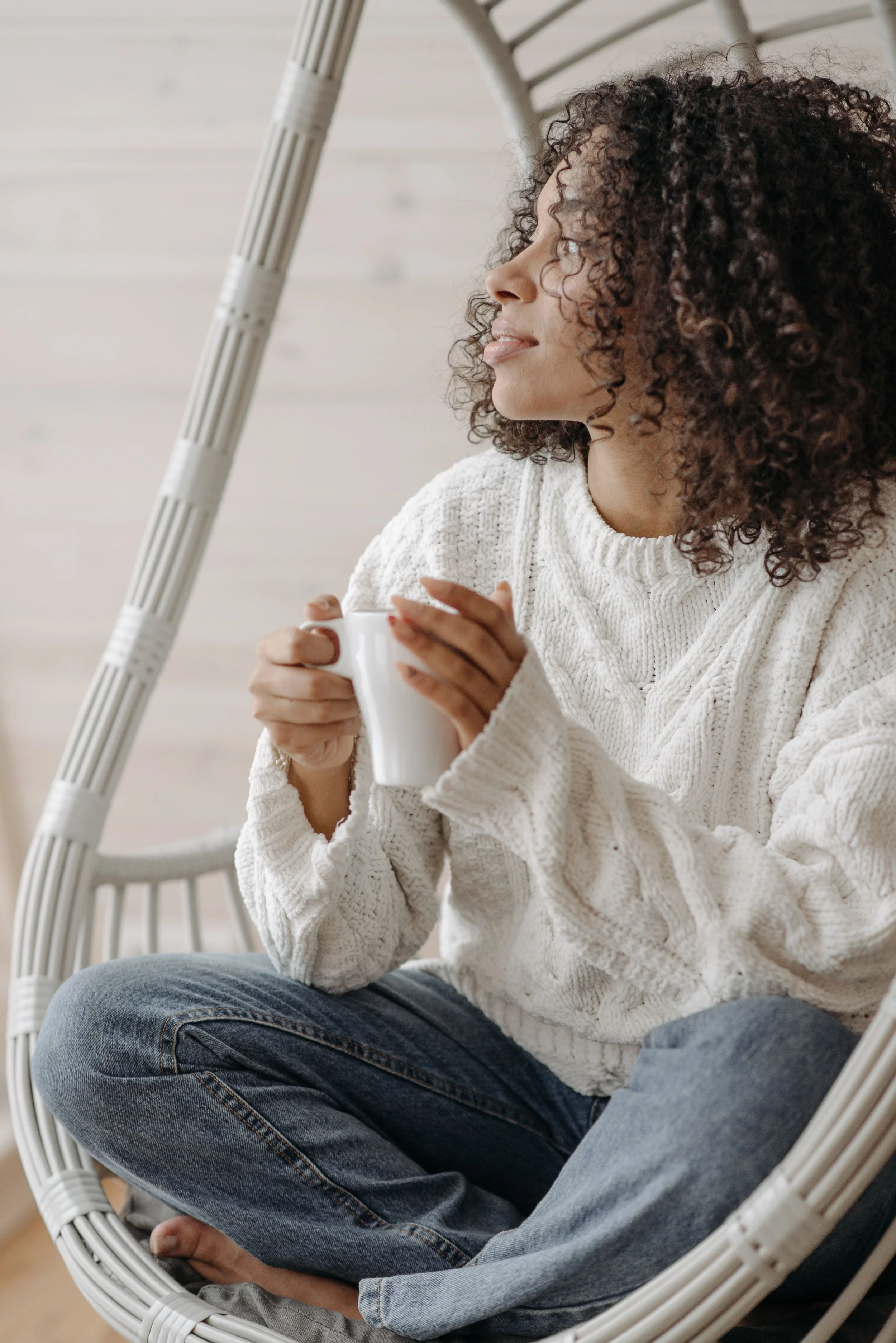 A woman with curly hair sitting in a hanging wicker chair, holding a white mug, wearing a white knitted sweater and jeans, looking to the side in a cozy indoor setting.