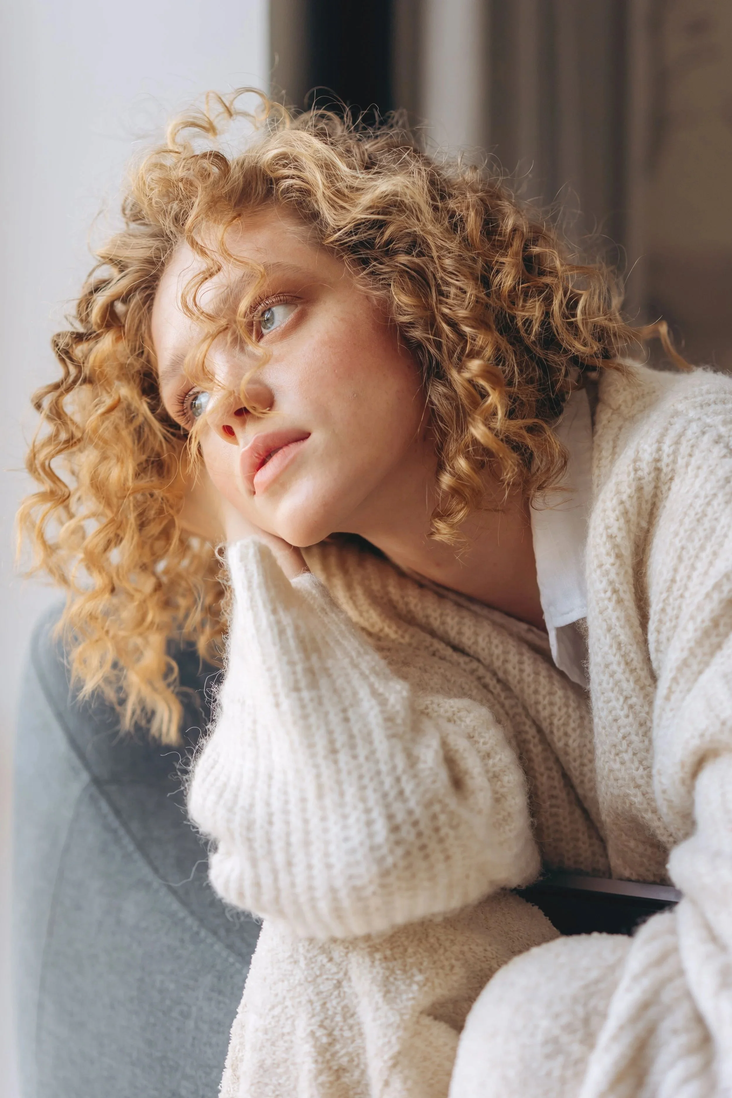 A woman with curly blonde hair and blue eyes, wearing a beige sweater, looking out of a window.