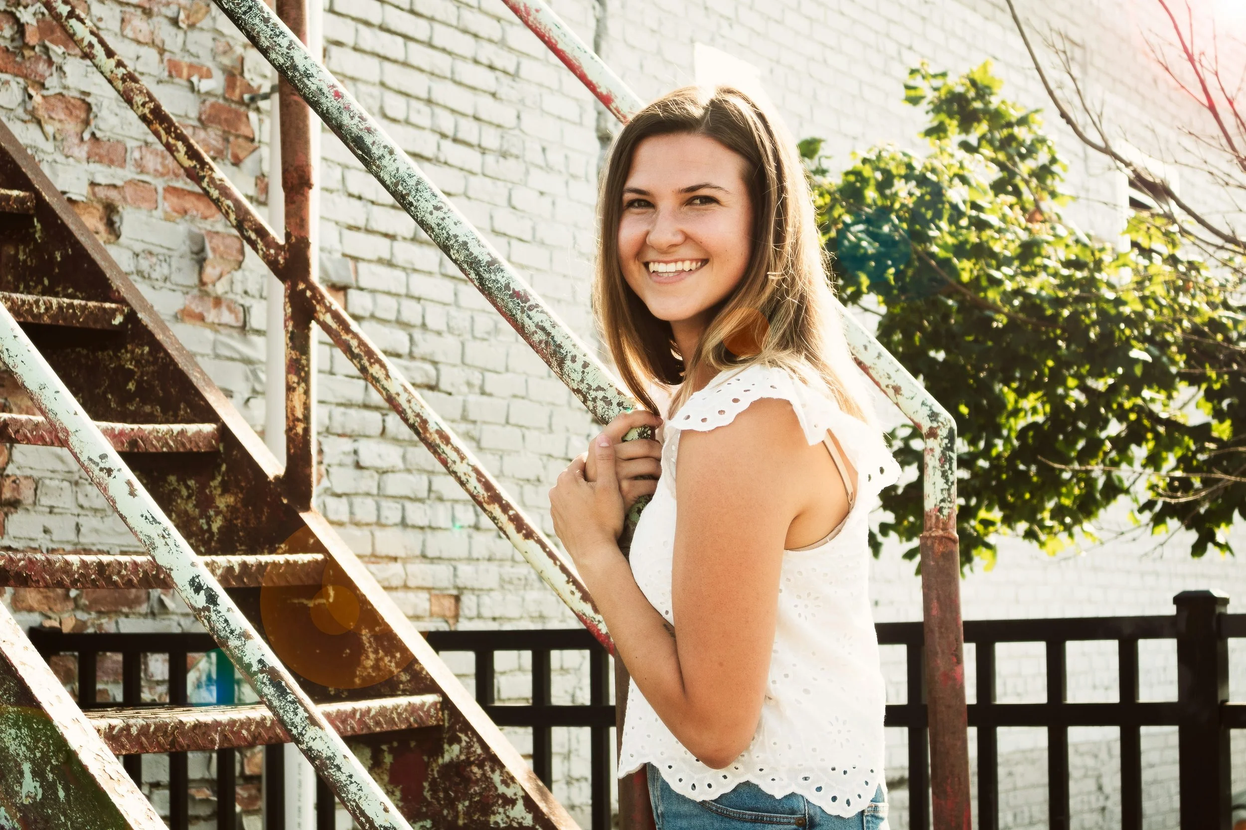 A young woman with long brown hair smiling outdoors near a rusty staircase and white brick wall, wearing a white eyelet sleeveless top and blue jeans.