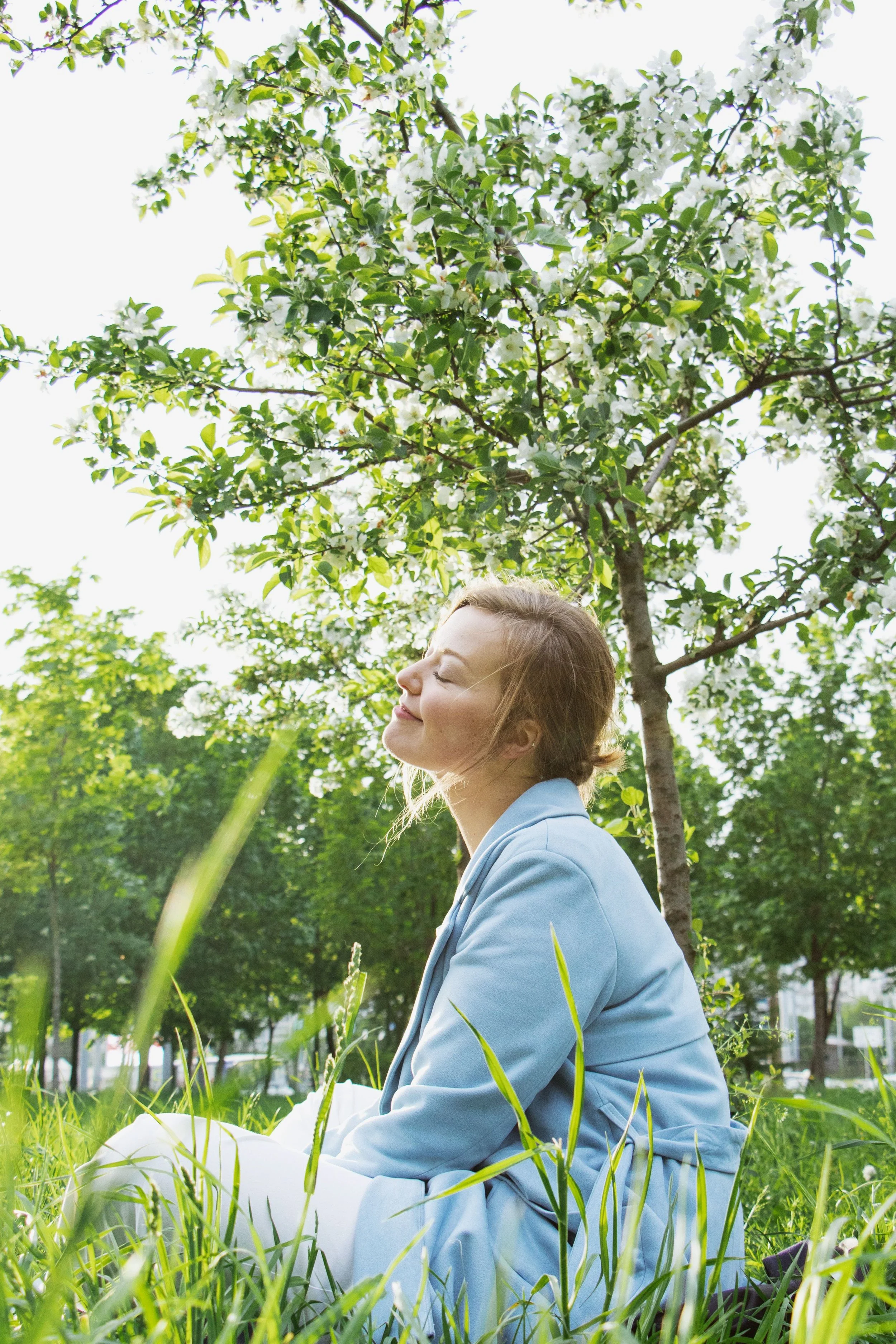 A young woman with brown hair sitting cross-legged on the grass in a park, smiling with her eyes closed under a blooming tree with white flowers.
