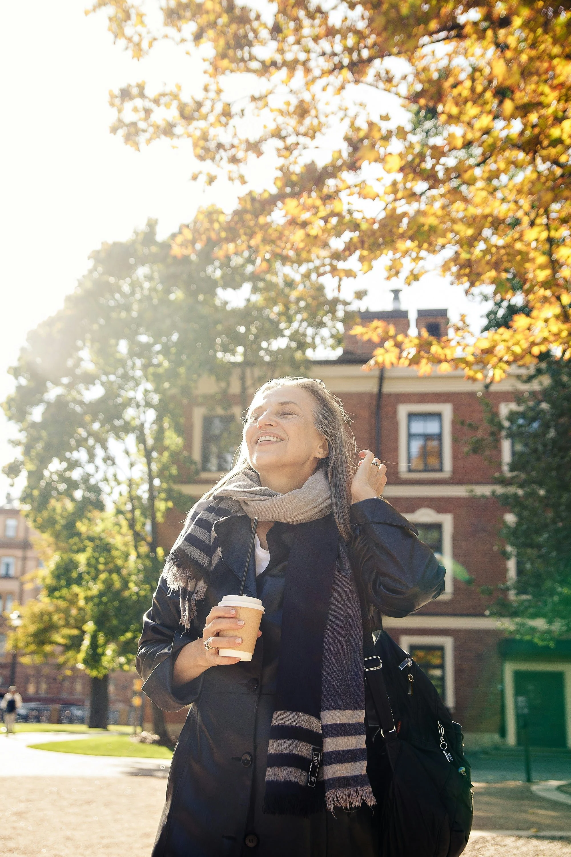 A woman smiling and holding a coffee cup outdoors in front of autumn trees and a brick building.