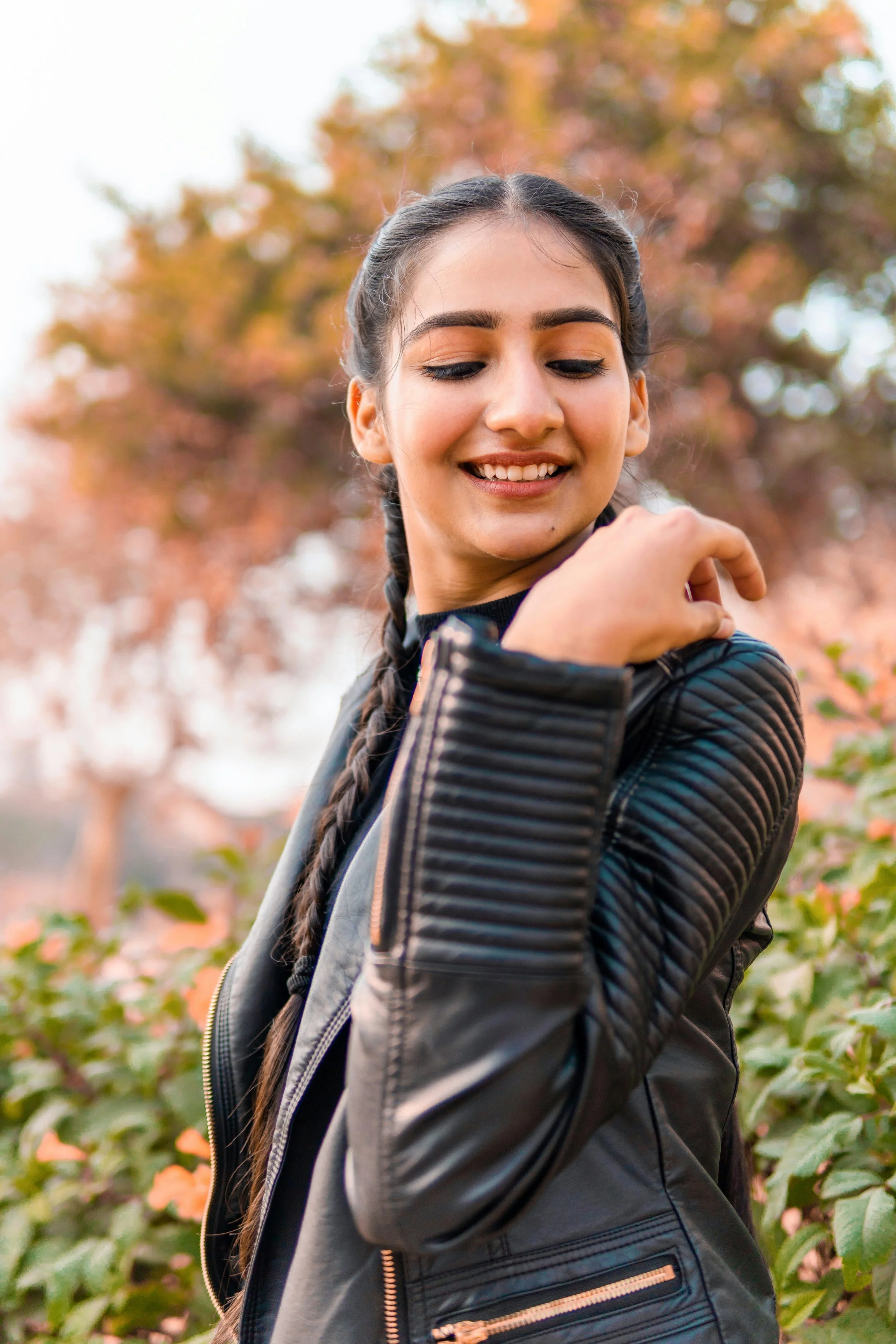 A young woman with long braided hair, closed eyes, smiling, and wearing a black leather jacket, standing outdoors with fall foliage in the background.