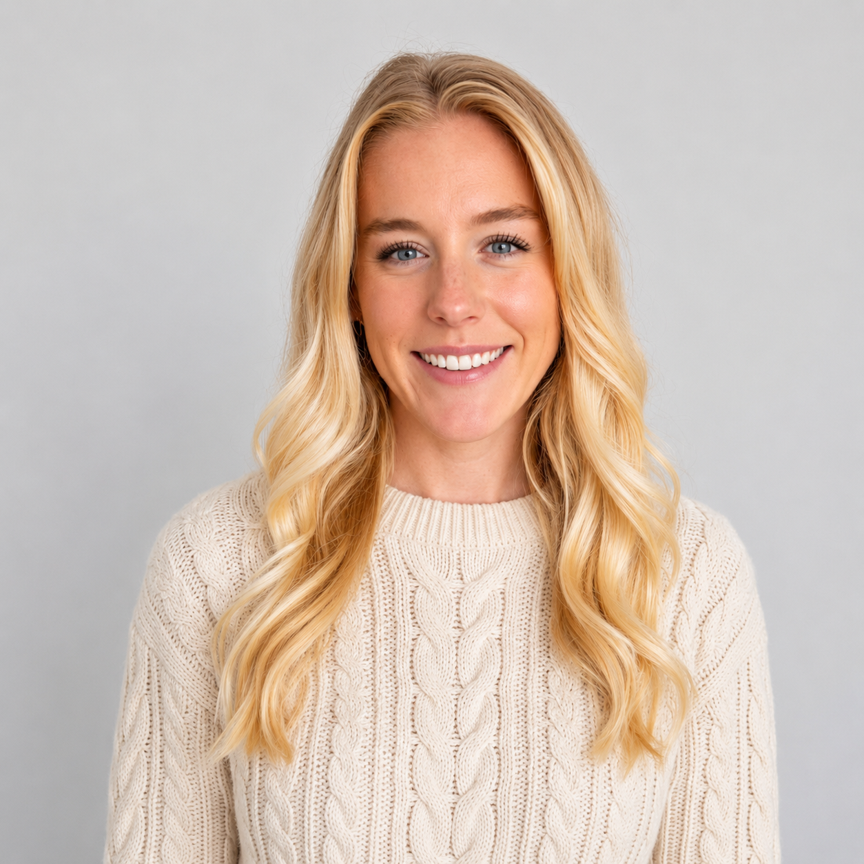 A smiling woman with blonde hair, wearing a cream cable knit sweater, against a light gray background.