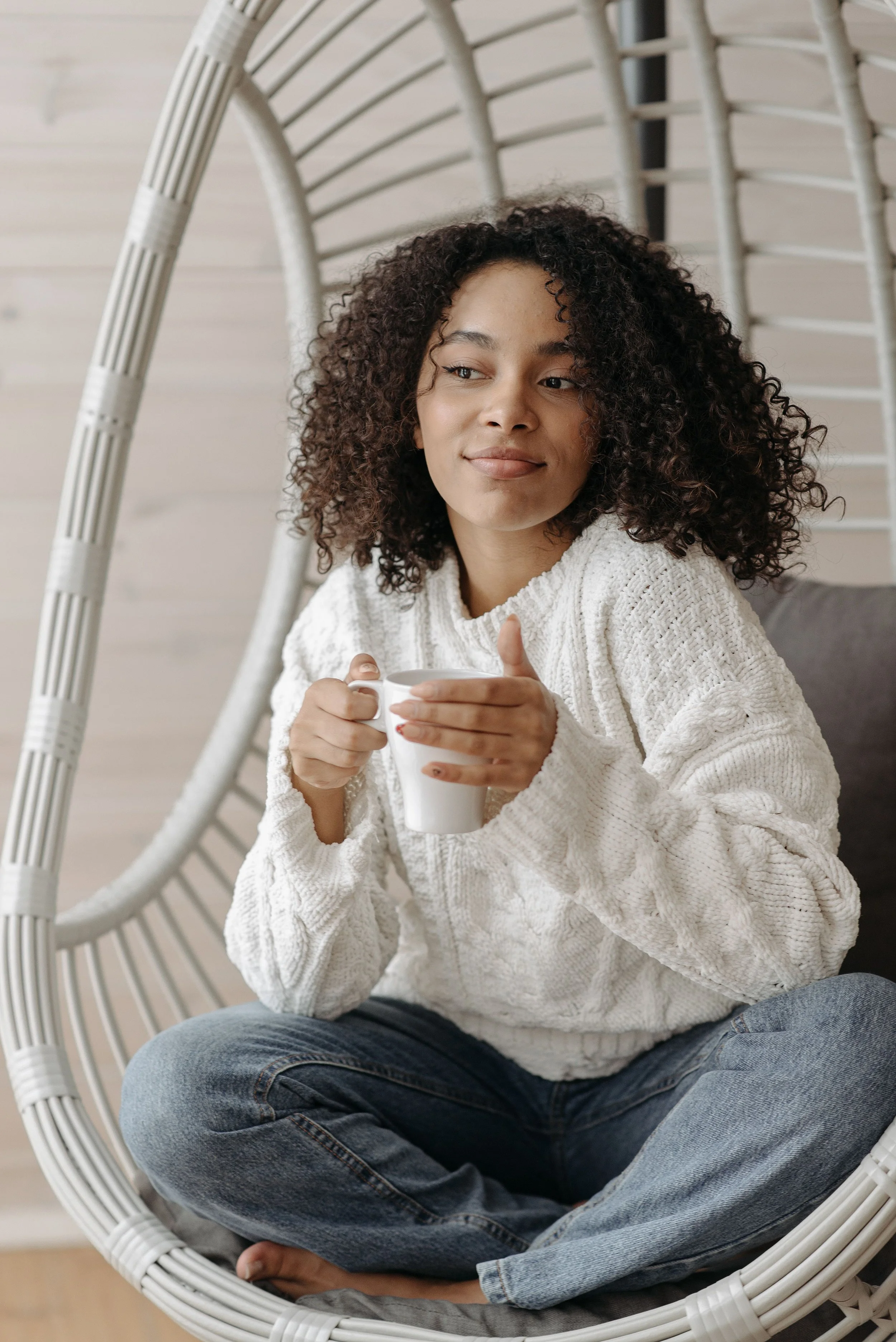 A young woman with curly hair sitting cross-legged in a white wicker hanging chair, holding a white mug, smiling softly, wearing a cream sweater and blue jeans, in a room with light-colored wooden wall panels.