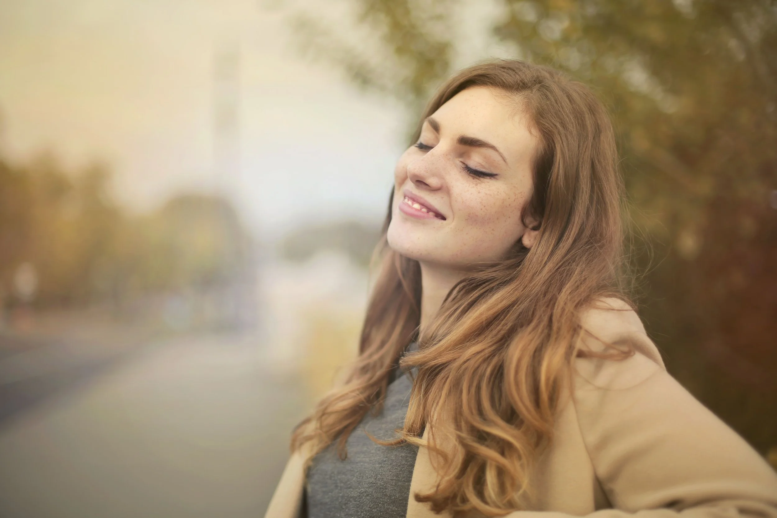 A young woman with long, wavy red hair with freckles and closed eyes, smiling gently outdoors during daytime, with blurred autumn trees in the background.