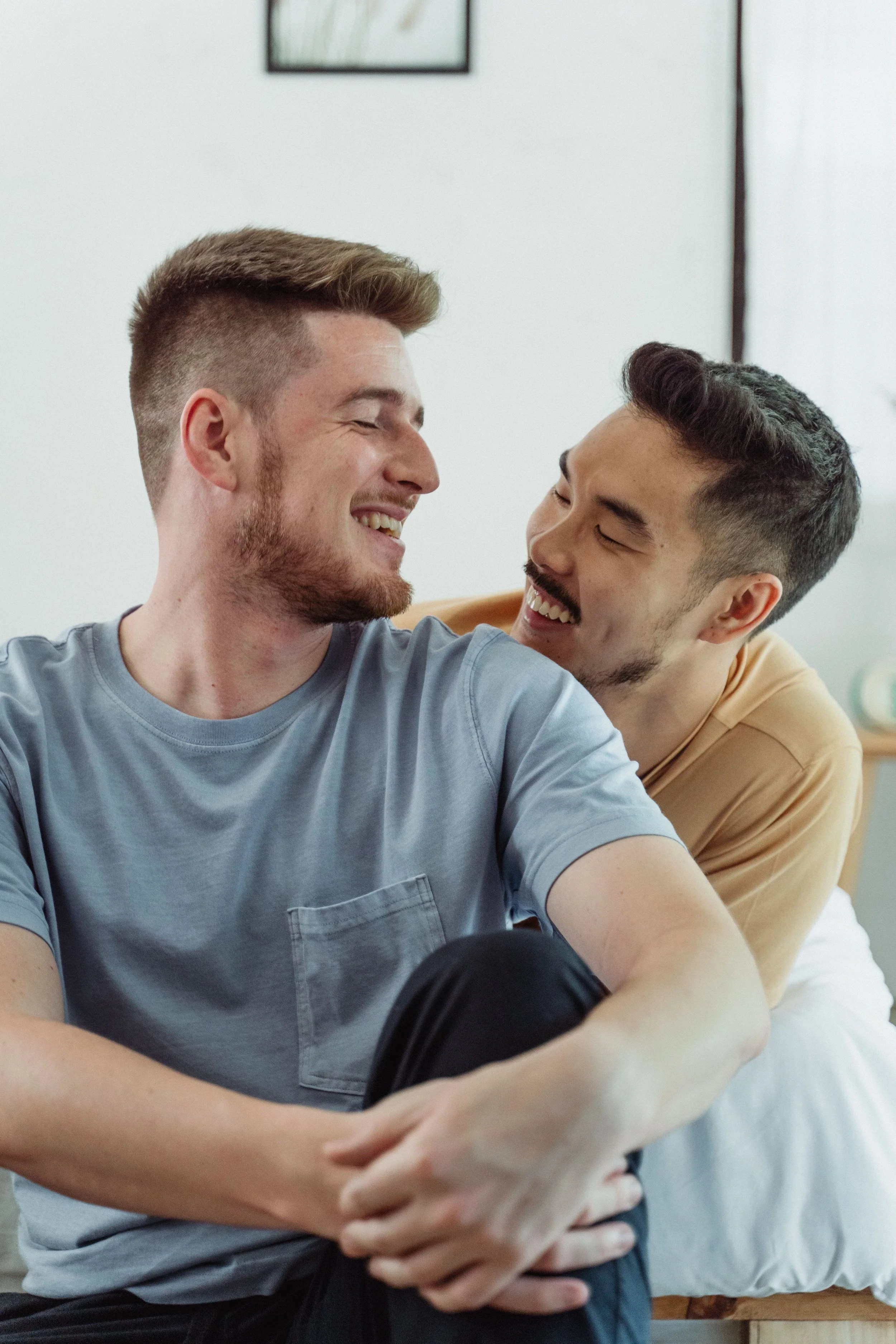 Two young men are sitting close together inside a room, looking at each other and smiling warmly. One is wearing a gray t-shirt, and the other is in a tan shirt. They seem happy and engaged.