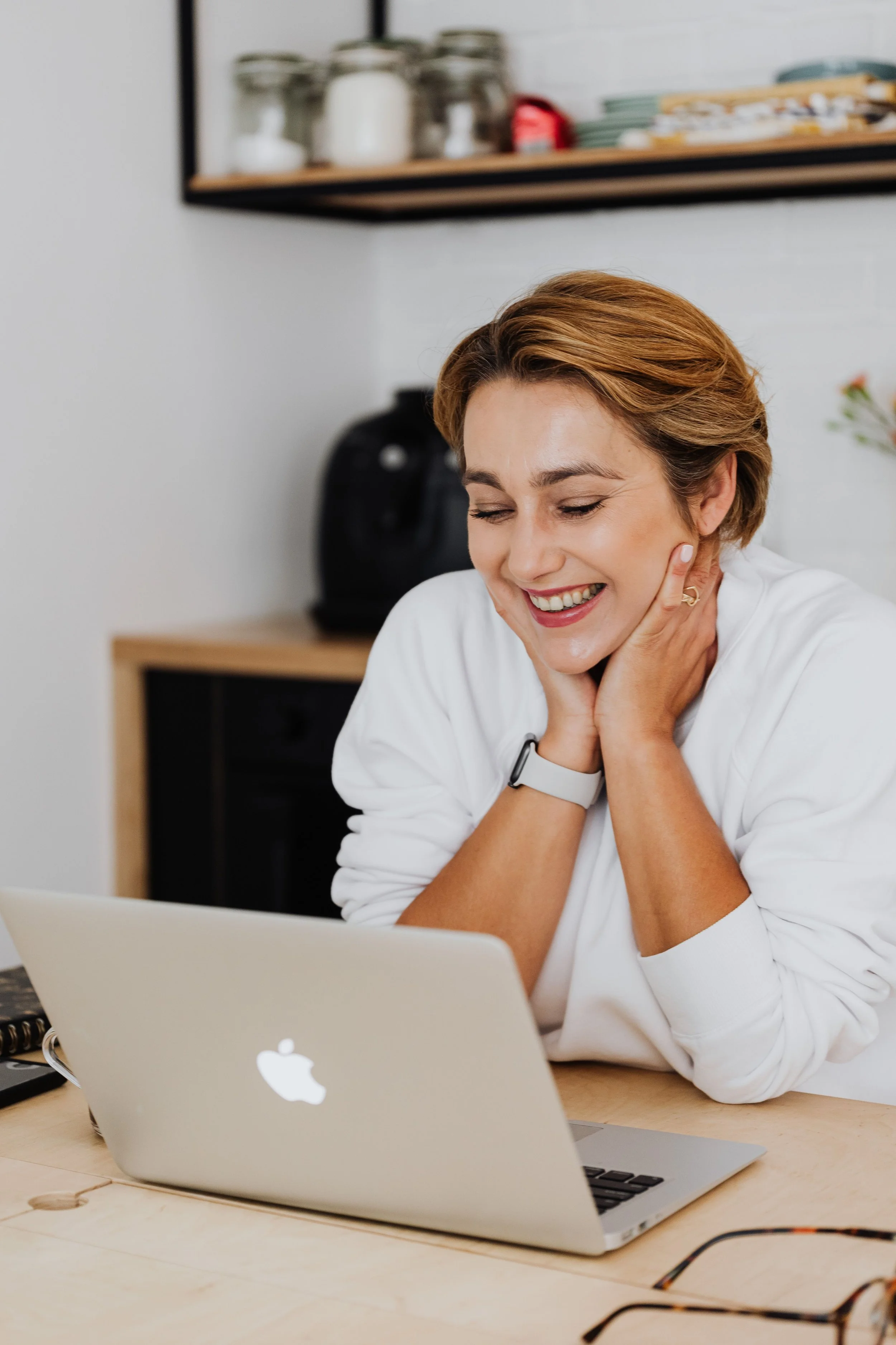 A woman with short, light brown hair smiling happily while sitting at a desk with an open MacBook in front of her. She is touching her neck with both hands. The background includes shelves with jars and plates, and a black appliance on a piece of furniture against a white wall.