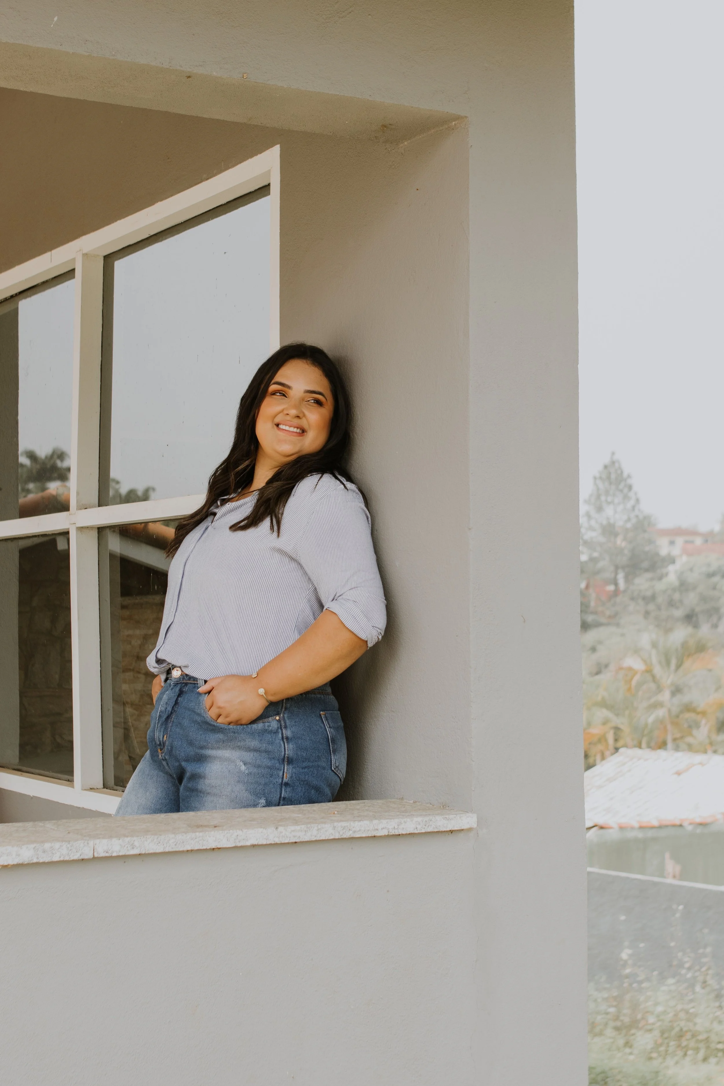 A young woman with long dark hair and a bright smile, wearing a light-colored blouse and jeans, leaning against an exterior wall beside a large window on a building. She is standing with her hand in her pocket, looking off to the side.