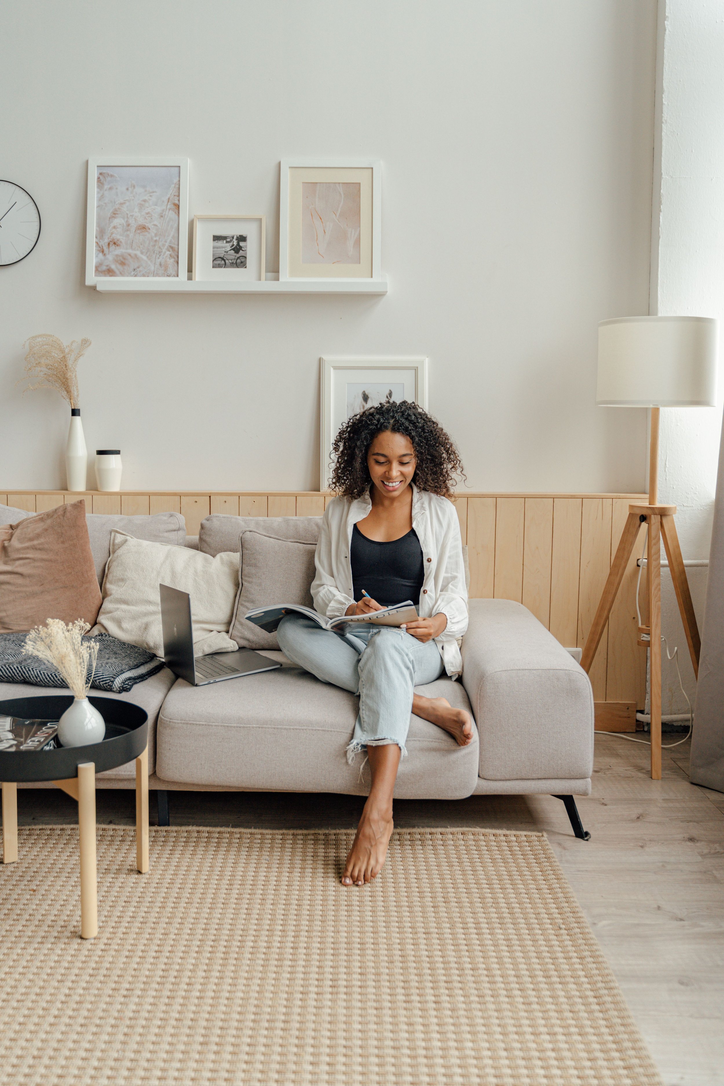 A woman sitting on a light-colored sofa in a cozy, modern living room, reading a magazine, with a laptop nearby. The room has neutral tones, simple decor, framed artwork on the white wall, a tall floor lamp, two white vases with dried plants, and a small round black tray table with decorative items. The woman is barefoot, wearing a black top and light jeans, with curly hair and smiling.
