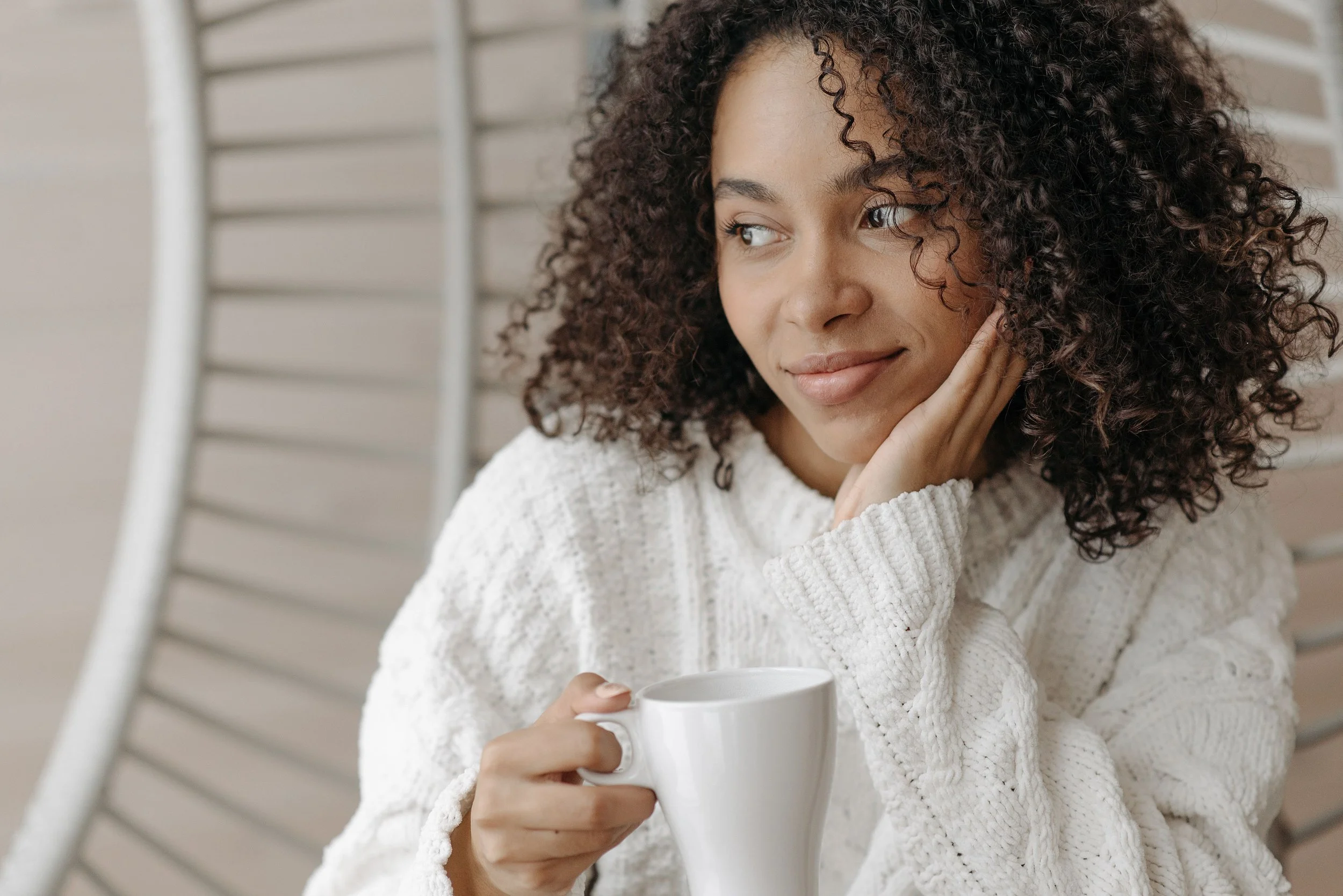 A woman with curly hair holding a white mug, wearing a cream sweater, sitting by a window with horizontal blinds.