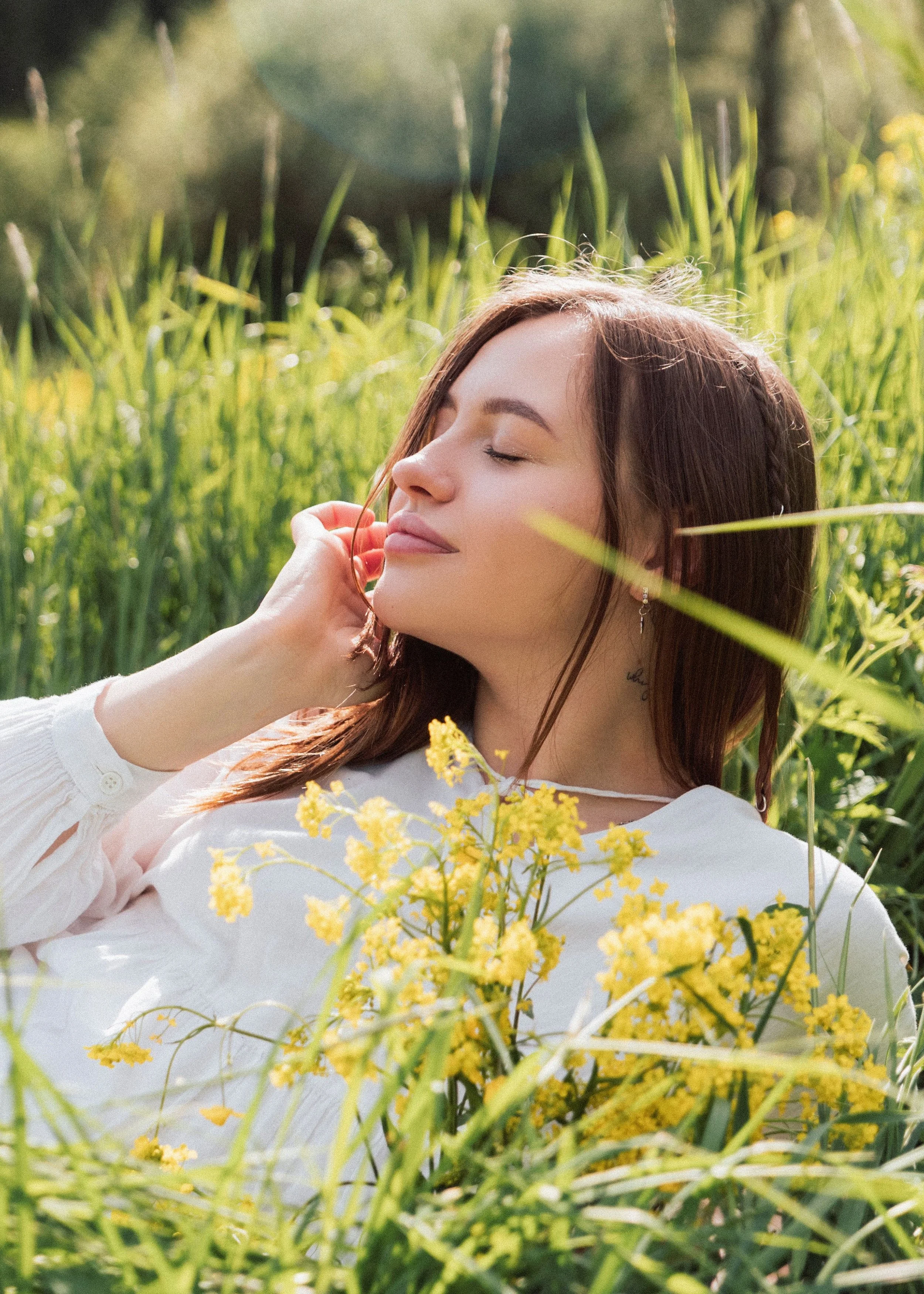 A young woman with long brown hair and light skin is lying in a field of tall green grass and yellow flowers, with her eyes closed and a peaceful expression on her face.
