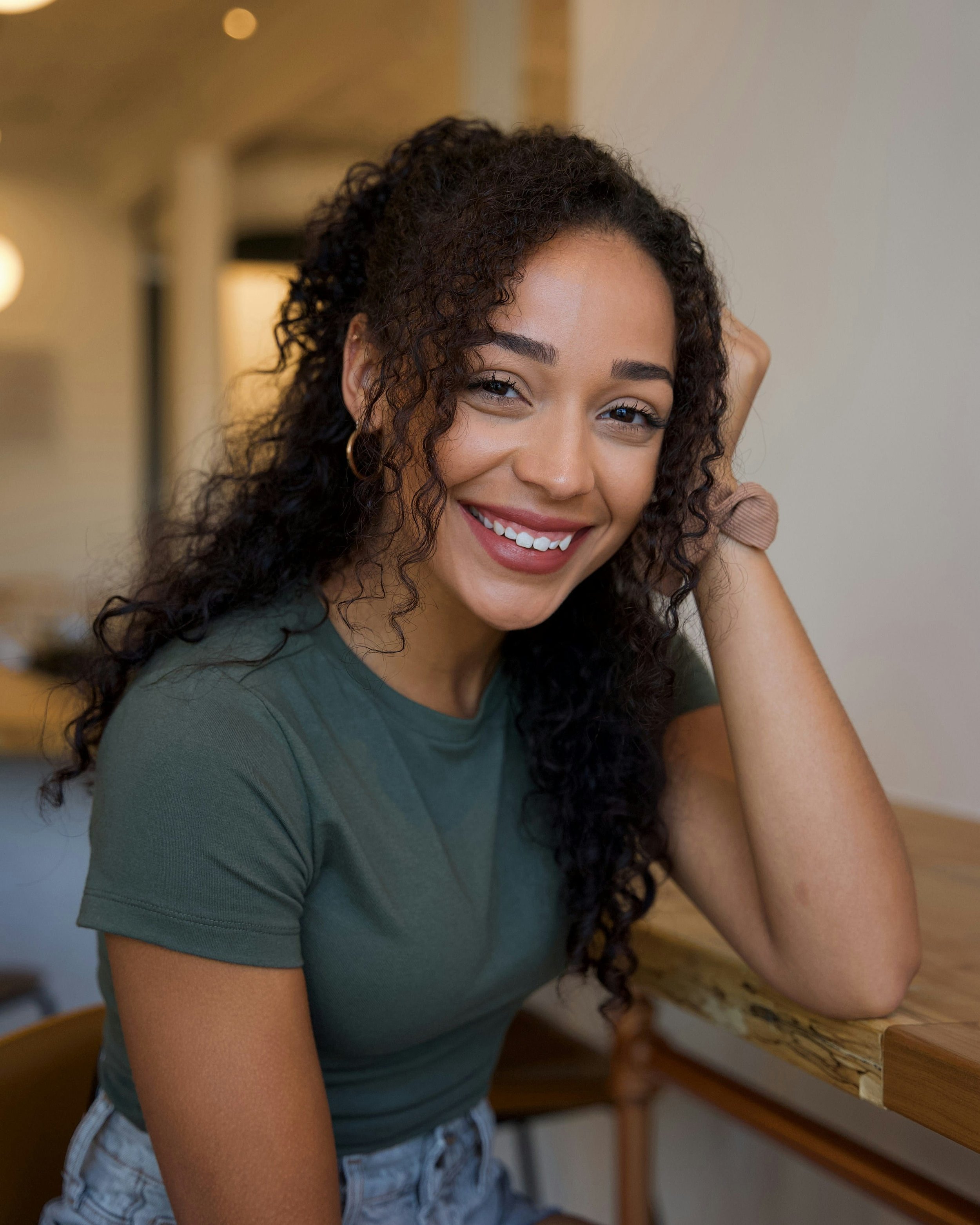A young woman with curly dark hair and a big smile, wearing a green t-shirt and light blue ripped jeans, sitting at a wooden table.