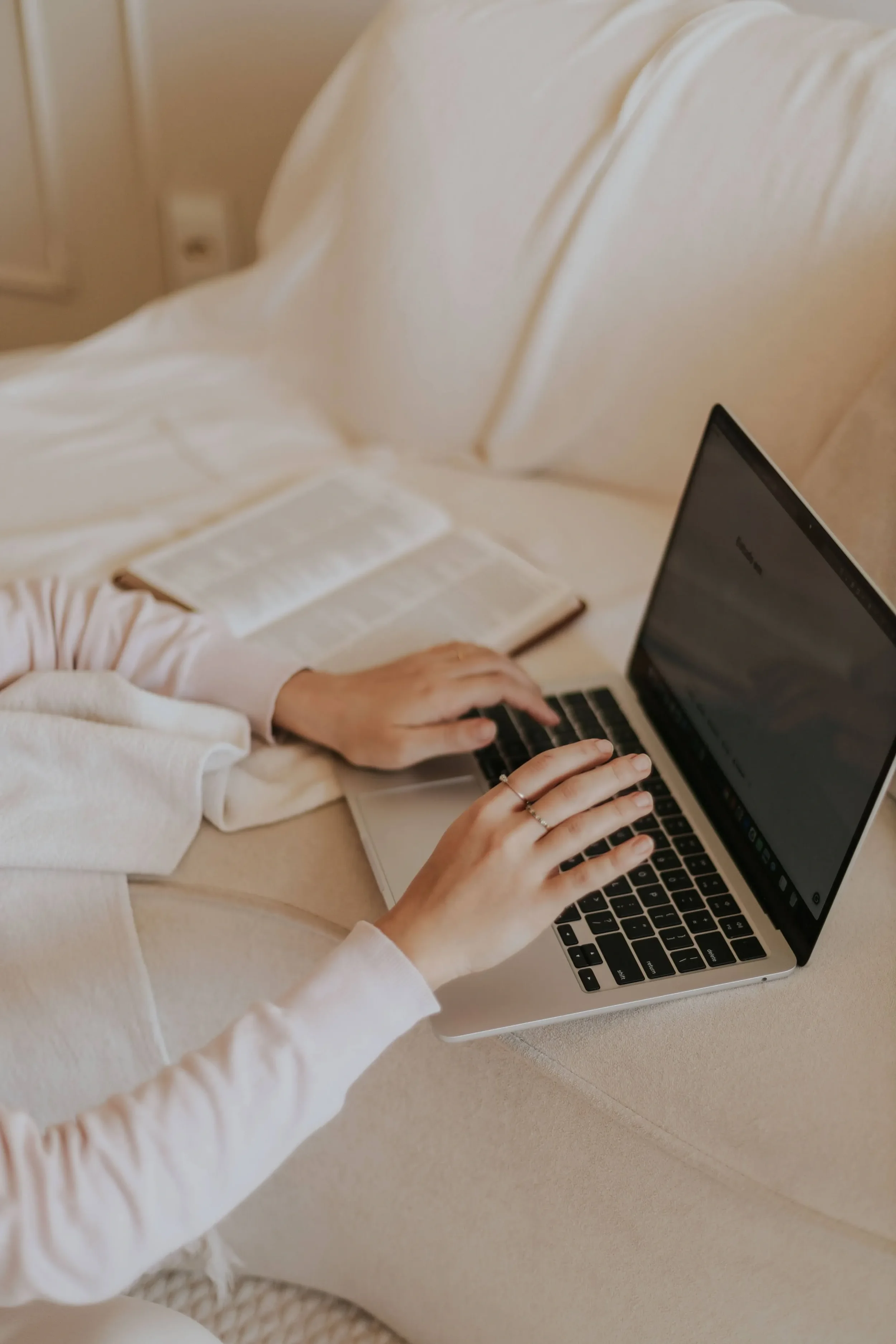 Person using a laptop on a beige sofa, wearing a light-colored long sleeve shirt, with a book nearby.