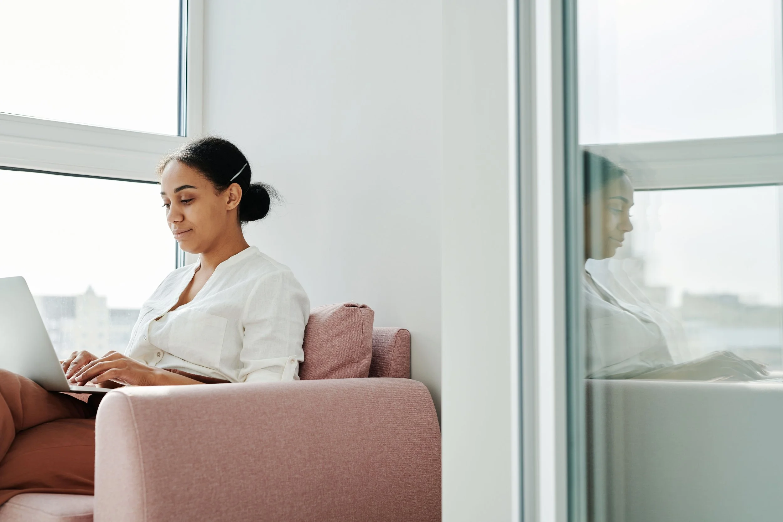 A woman with dark hair tied back sitting on a pink sofa, working on a laptop in a bright room with large windows.