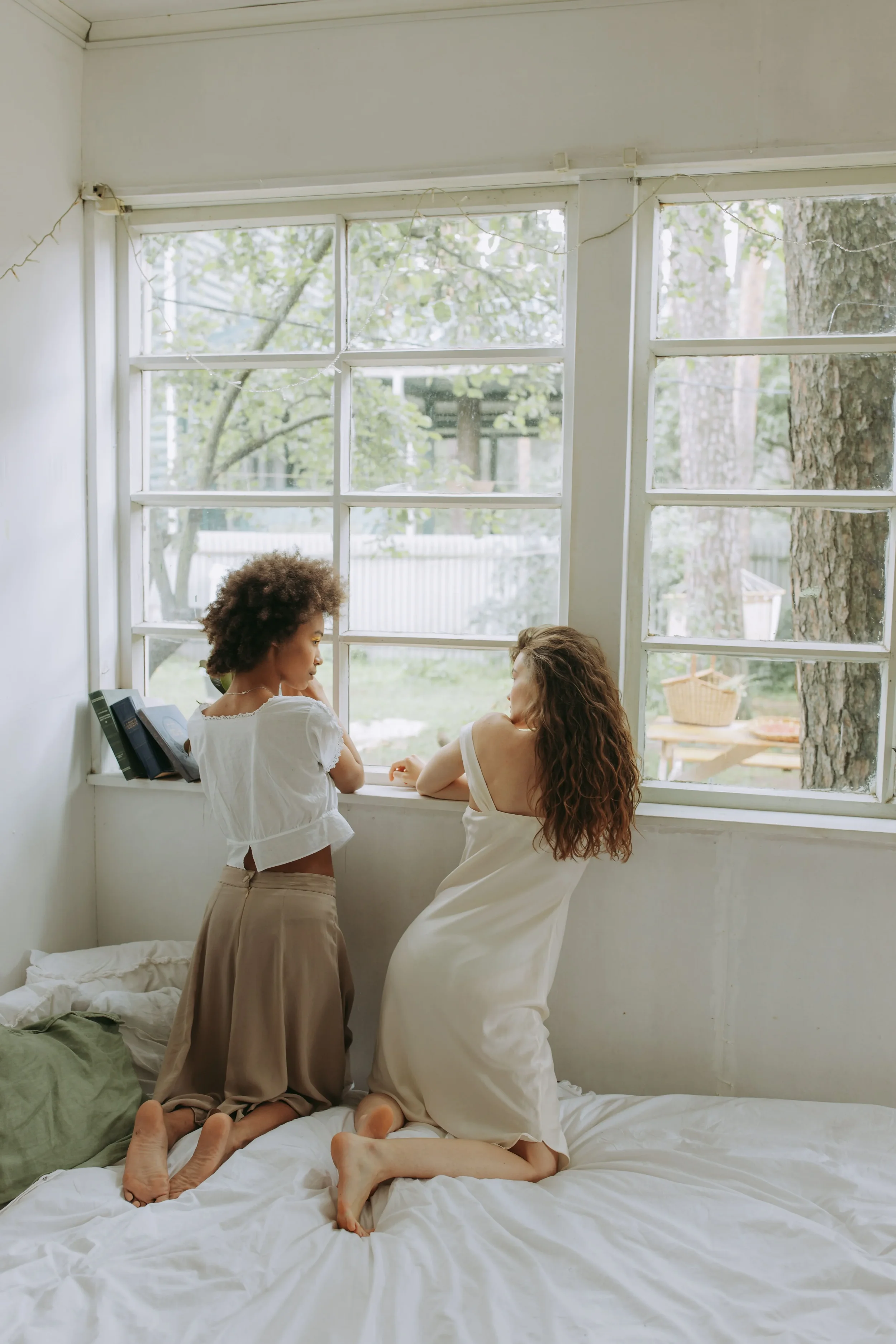 Two women are inside a bedroom with large windows and green trees outside, sitting on a bed with white sheets. One woman with curly hair is kneeling barefoot on the bed, wearing beige pants and a white top, looking at the other woman. The other woman has long wavy hair, wearing a white dress, and is kneeling on the bed, facing the woman with curly hair.