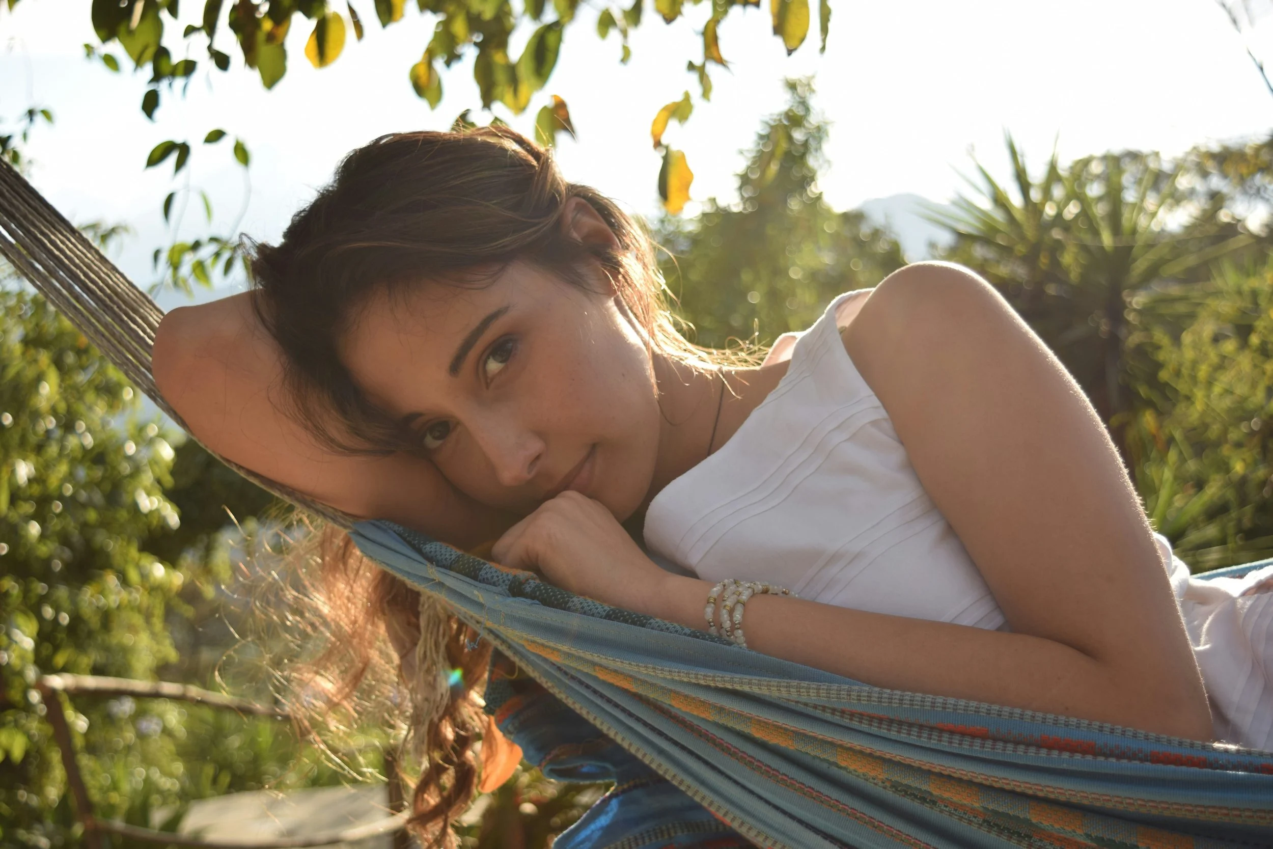 A woman with long brown hair lying in a hammock outdoors near green trees, with sunlight shining through the leaves.