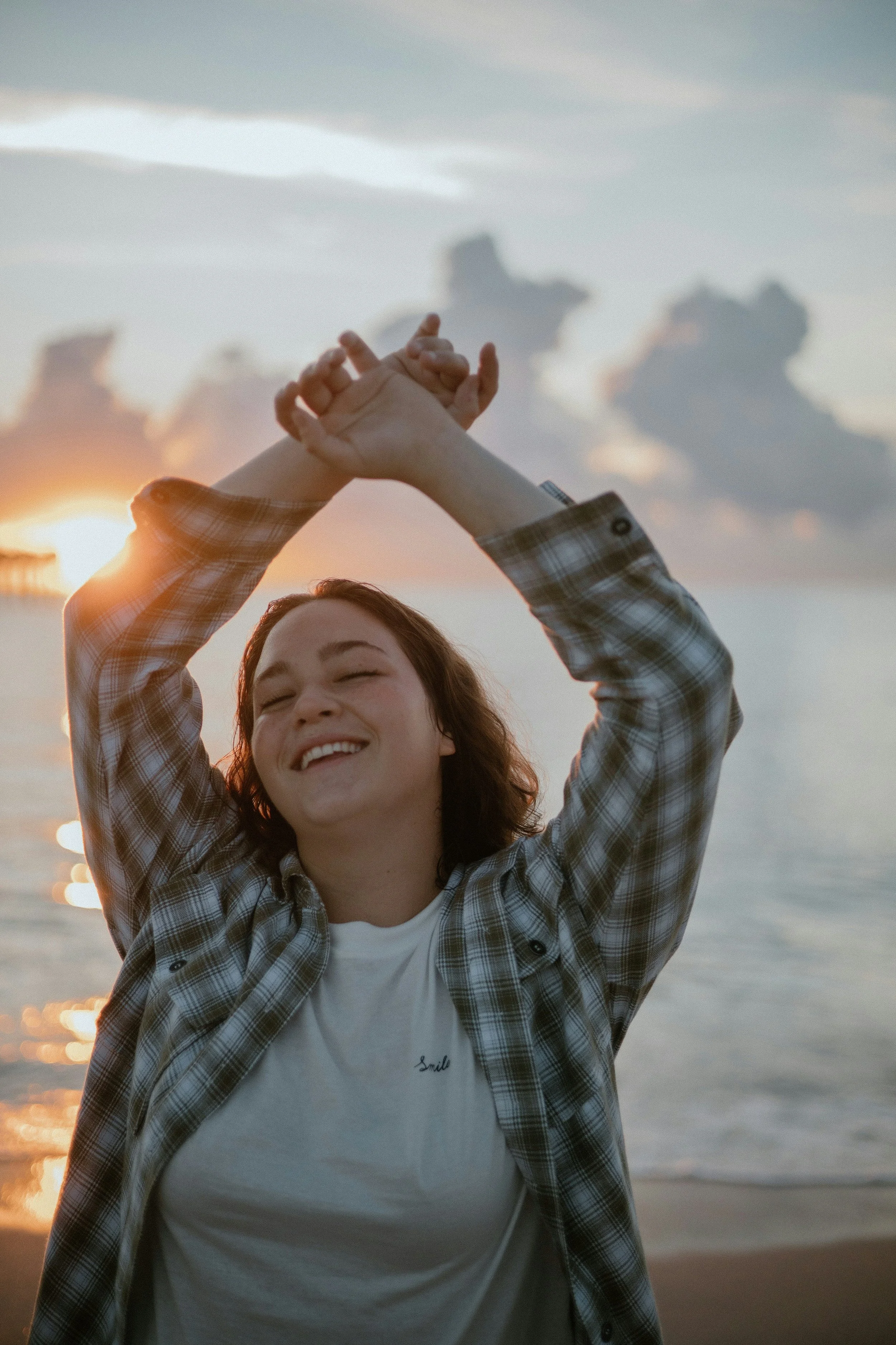 A young woman smiling with eyes closed, wearing a plaid shirt over a white t-shirt with the word 'Smile' on it, standing on a beach at sunset with her hands raised above her head, forming a heart shape.